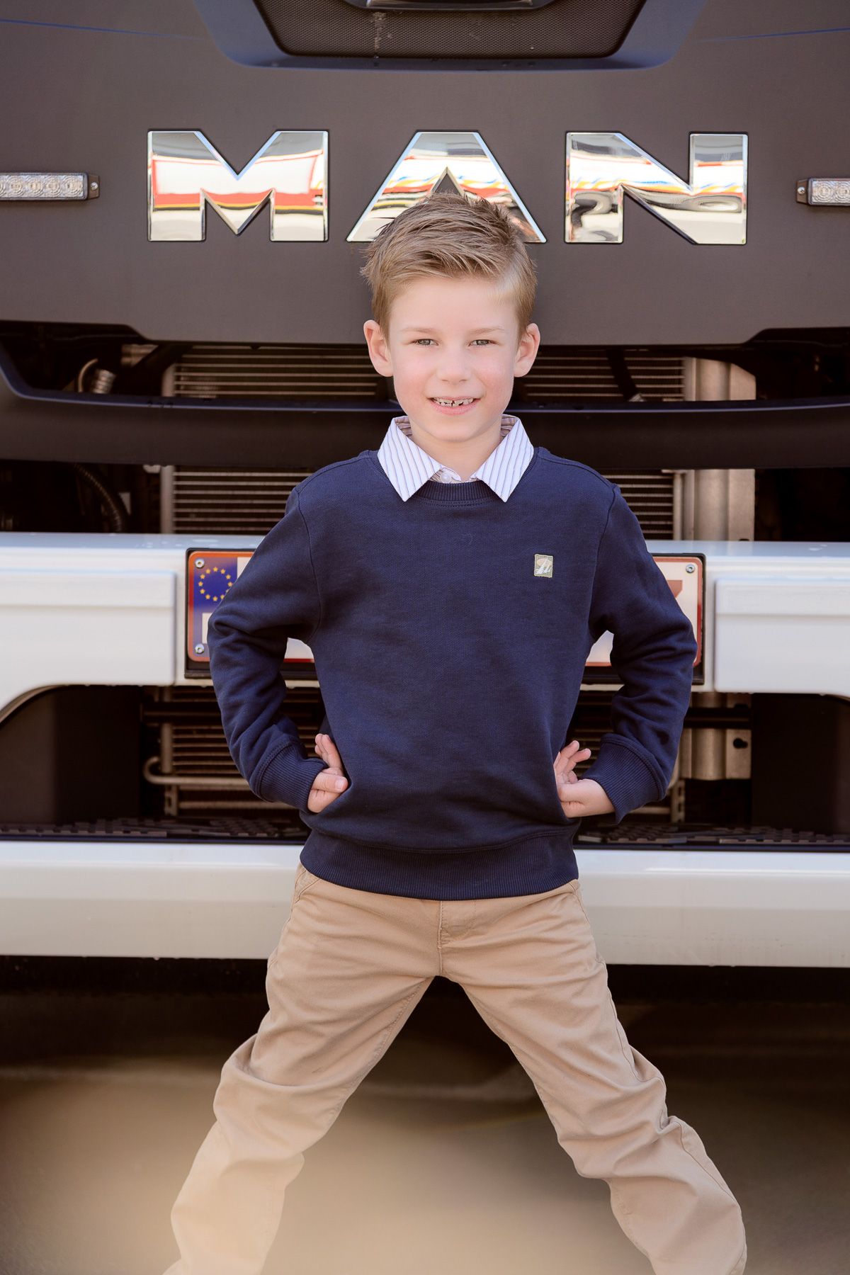 Young boy in navy sweater posing confidently in front of a MAN truck, smiling with hands on hips.