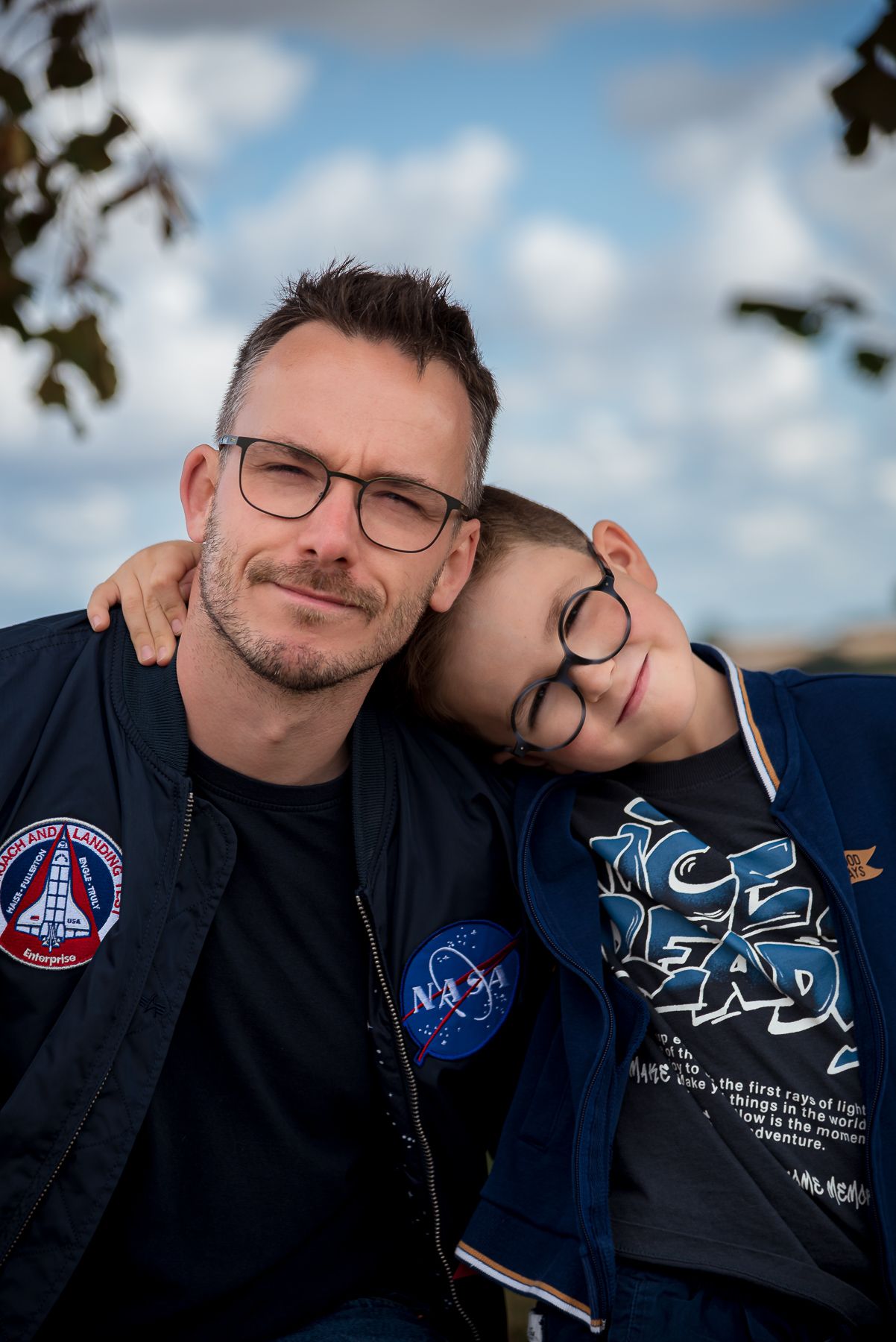 A father and son wearing matching navy jackets pose together outdoors under a cloudy sky.