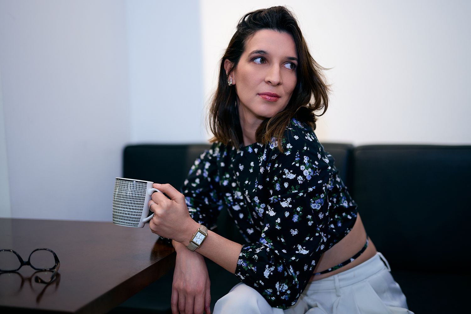 Young woman in floral blouse holding a coffee mug, sitting thoughtfully on a dark sofa indoors.