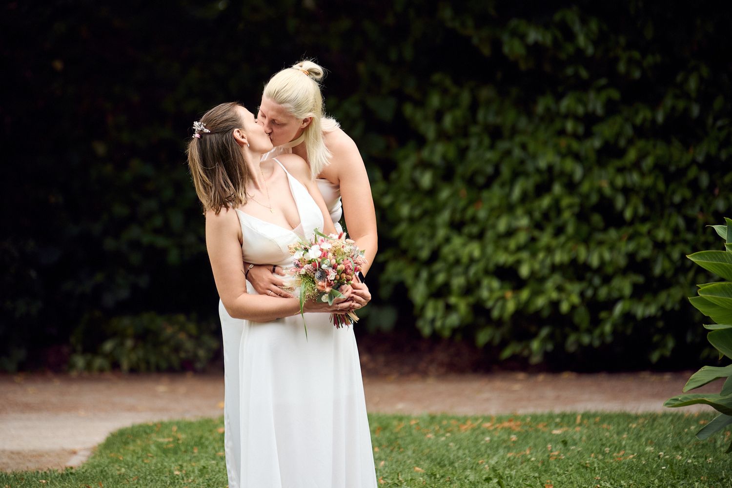 Two brides in white dresses share a kiss while holding a colorful bouquet in a lush garden setting.