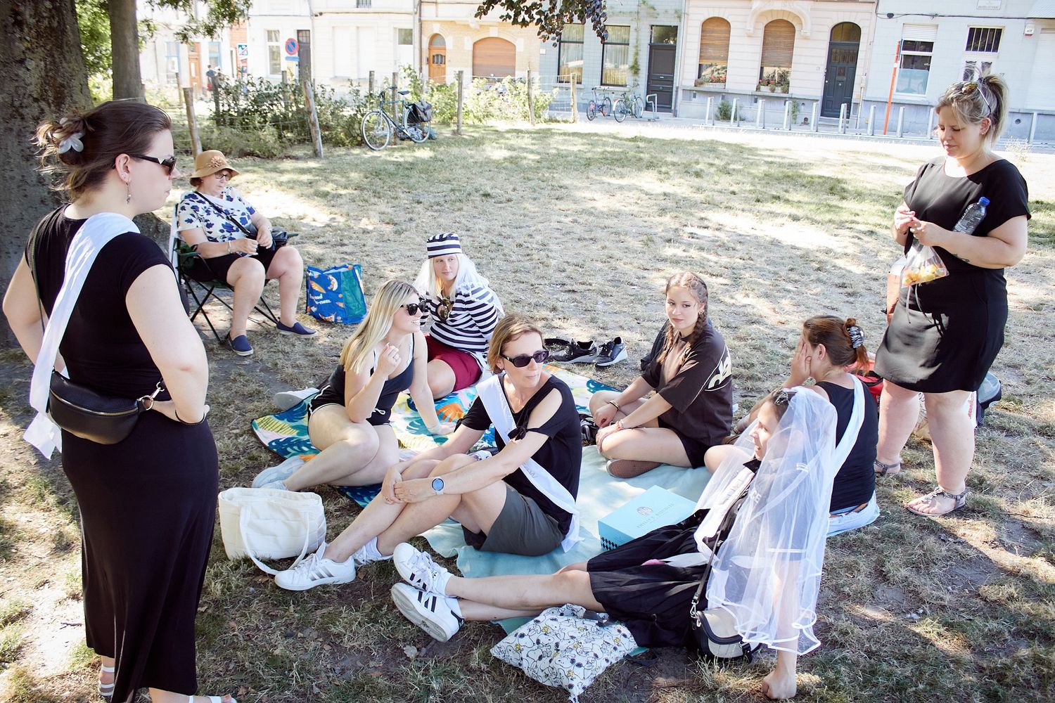 Group of women in black dresses gathered in a sunny park, sitting on blankets during an outdoor bachelorette party celebration.