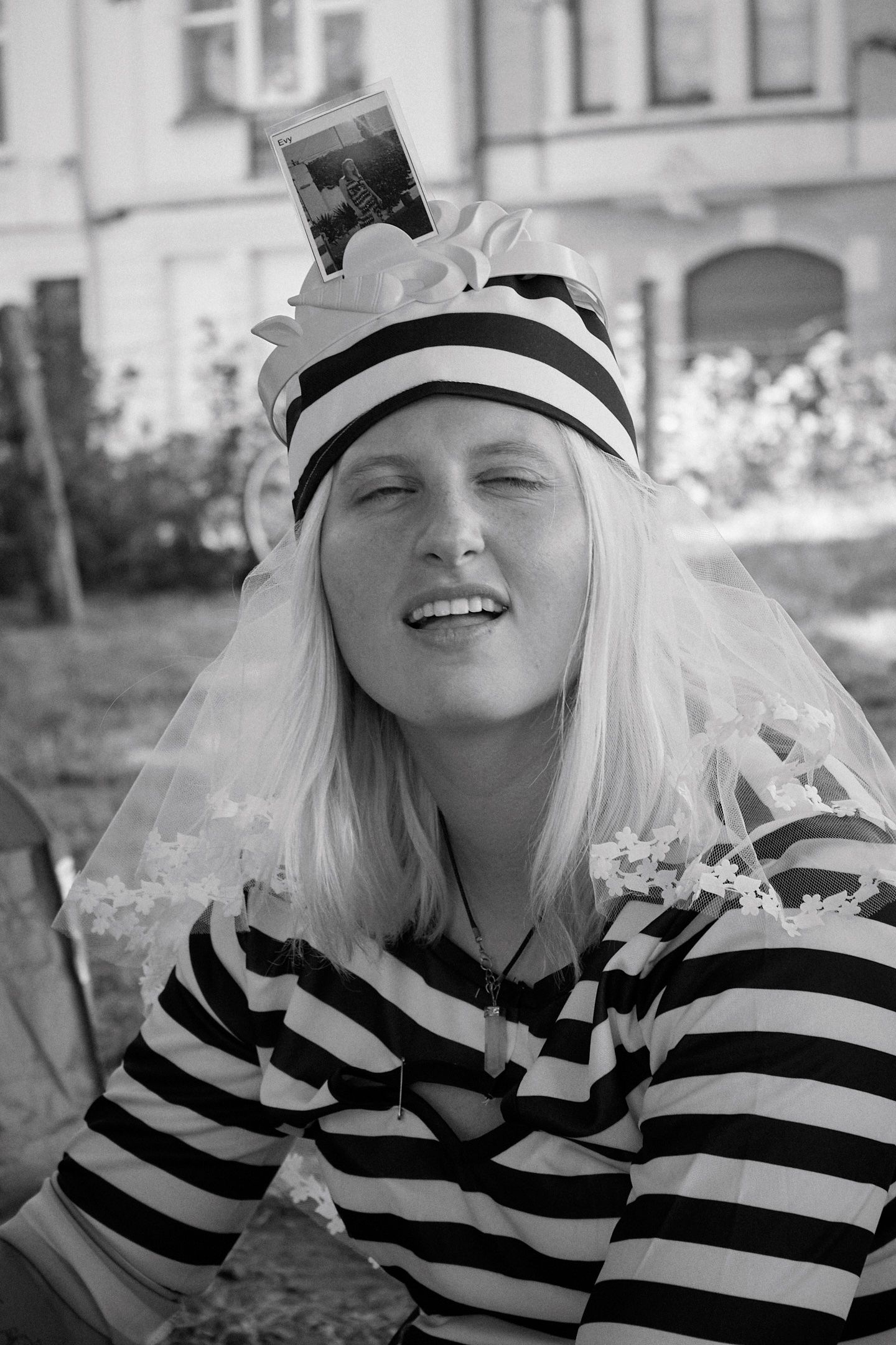 Black and white photo of a smiling blonde woman wearing a striped hat with a veil and striped shirt outdoors.