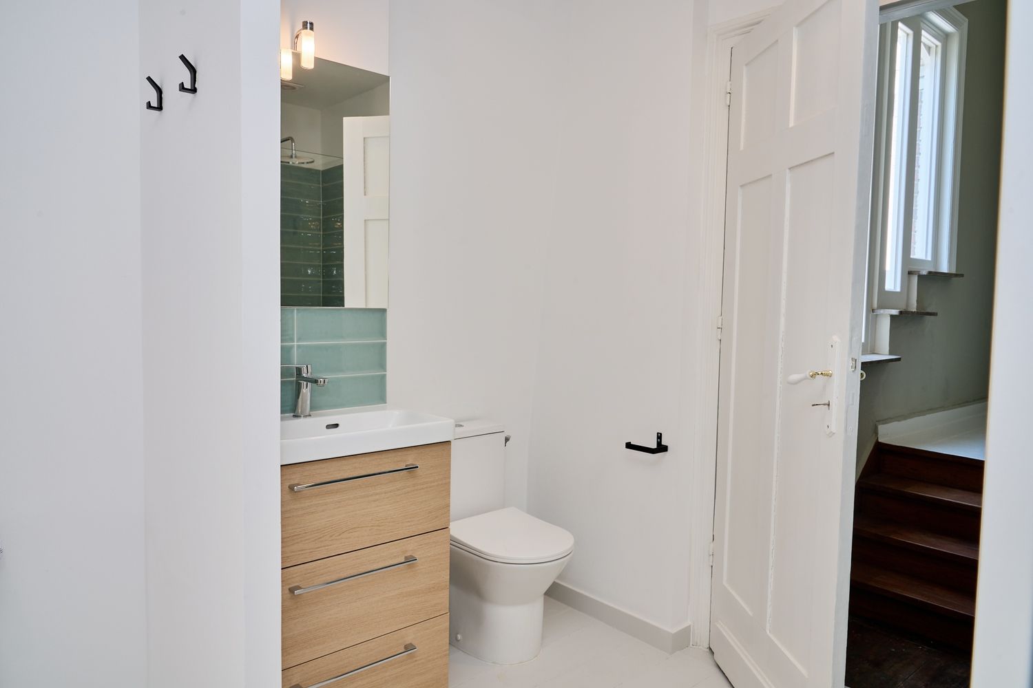 Modern bathroom with wood vanity, white toilet, green tile backsplash, and mirror cabinet next to a white door.