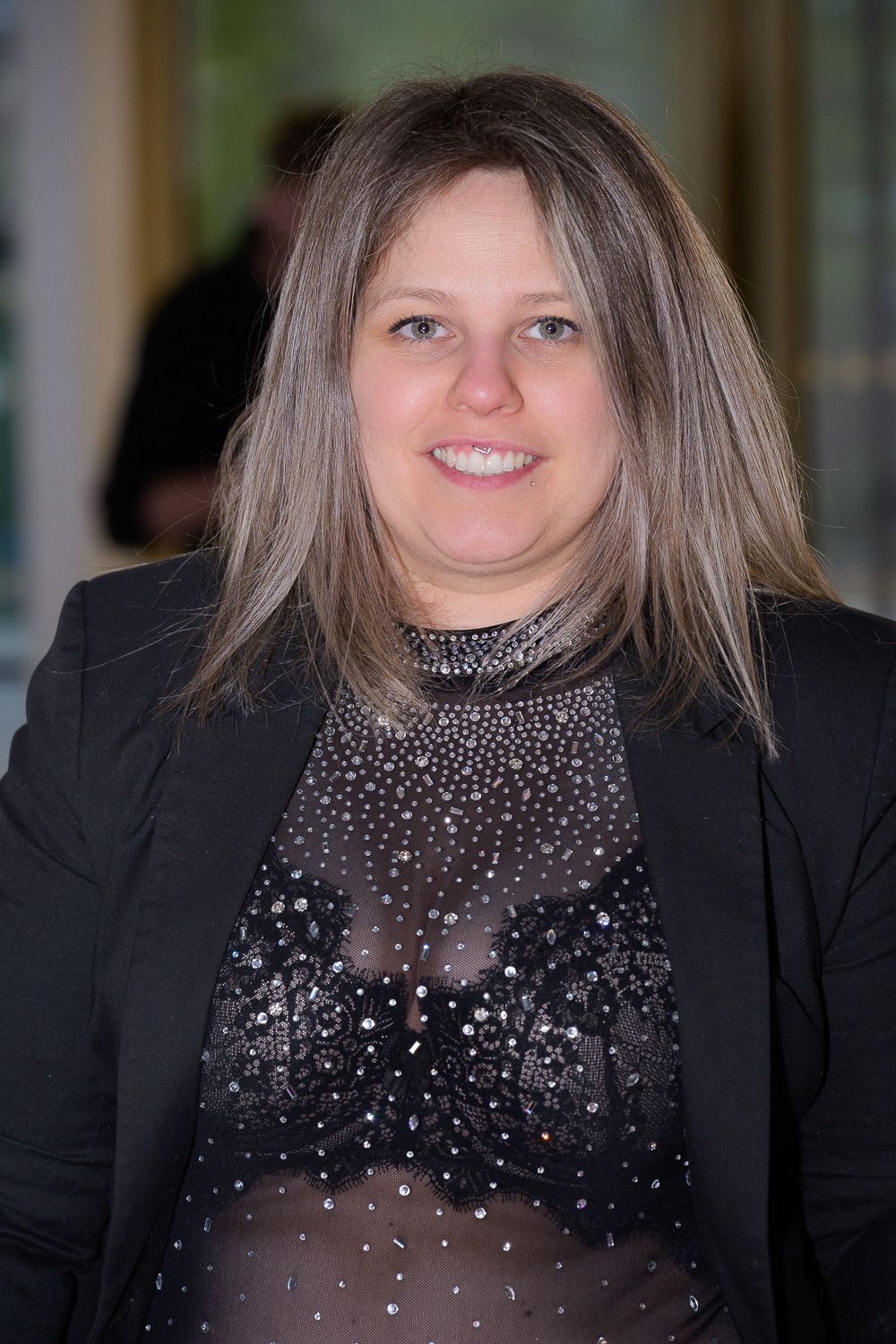 Smiling woman with long gray-blonde hair wearing a black blazer and sequined top at an indoor event.