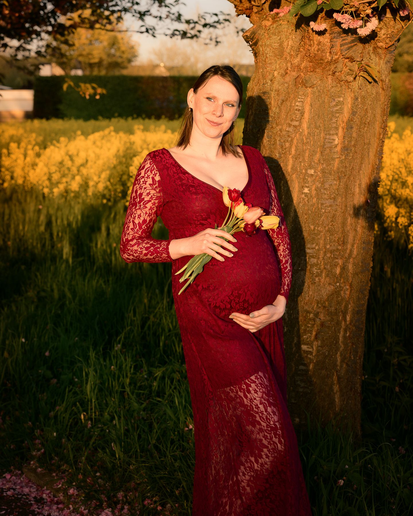 Pregnant woman in red lace gown holding yellow roses, leaning against tree in golden hour light with yellow flowers behind.