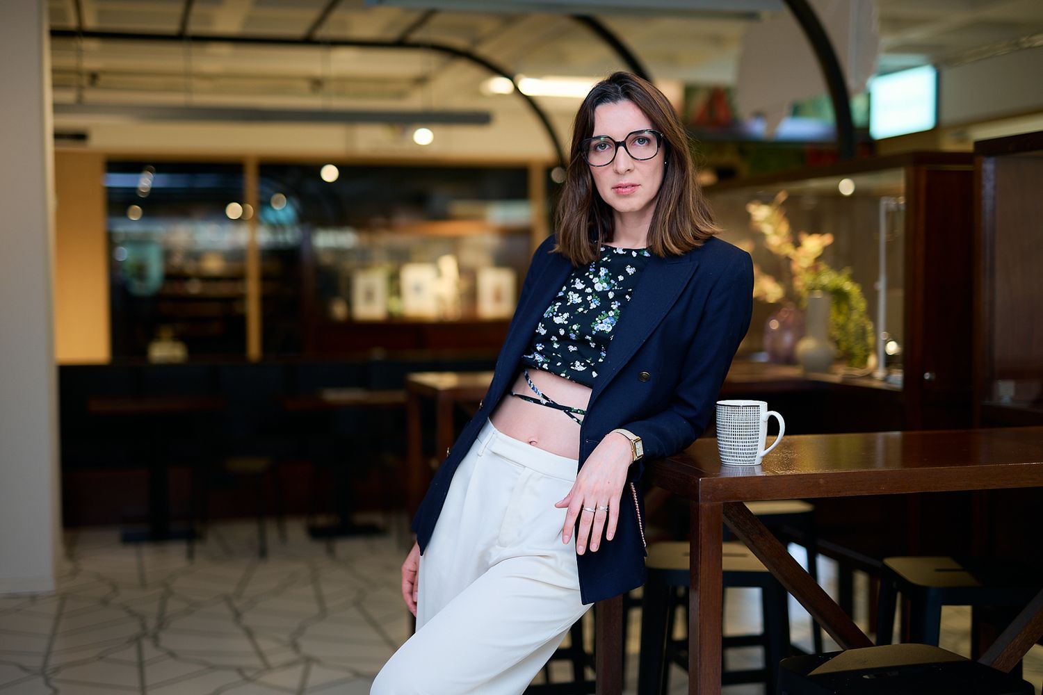 Young woman in navy blazer and white pants poses in modern cafe setting with glasses and floral top.