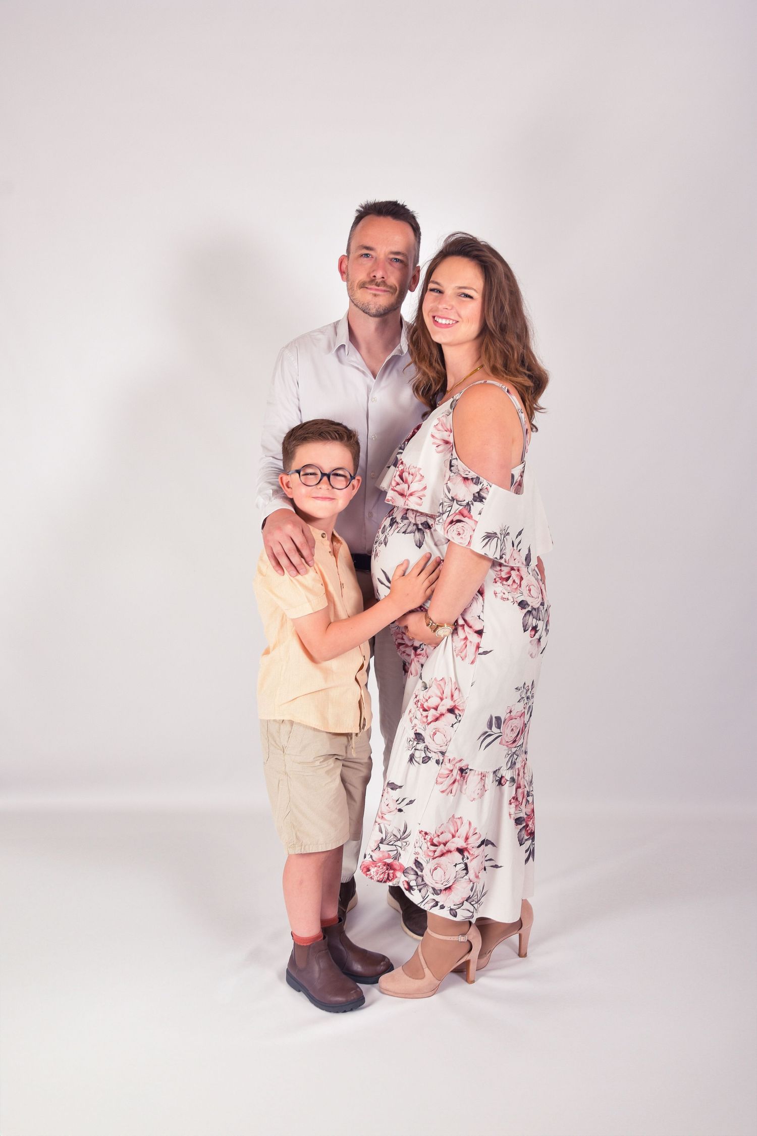 Smiling pregnant woman in floral dress posing with husband and young son in glasses in studio.