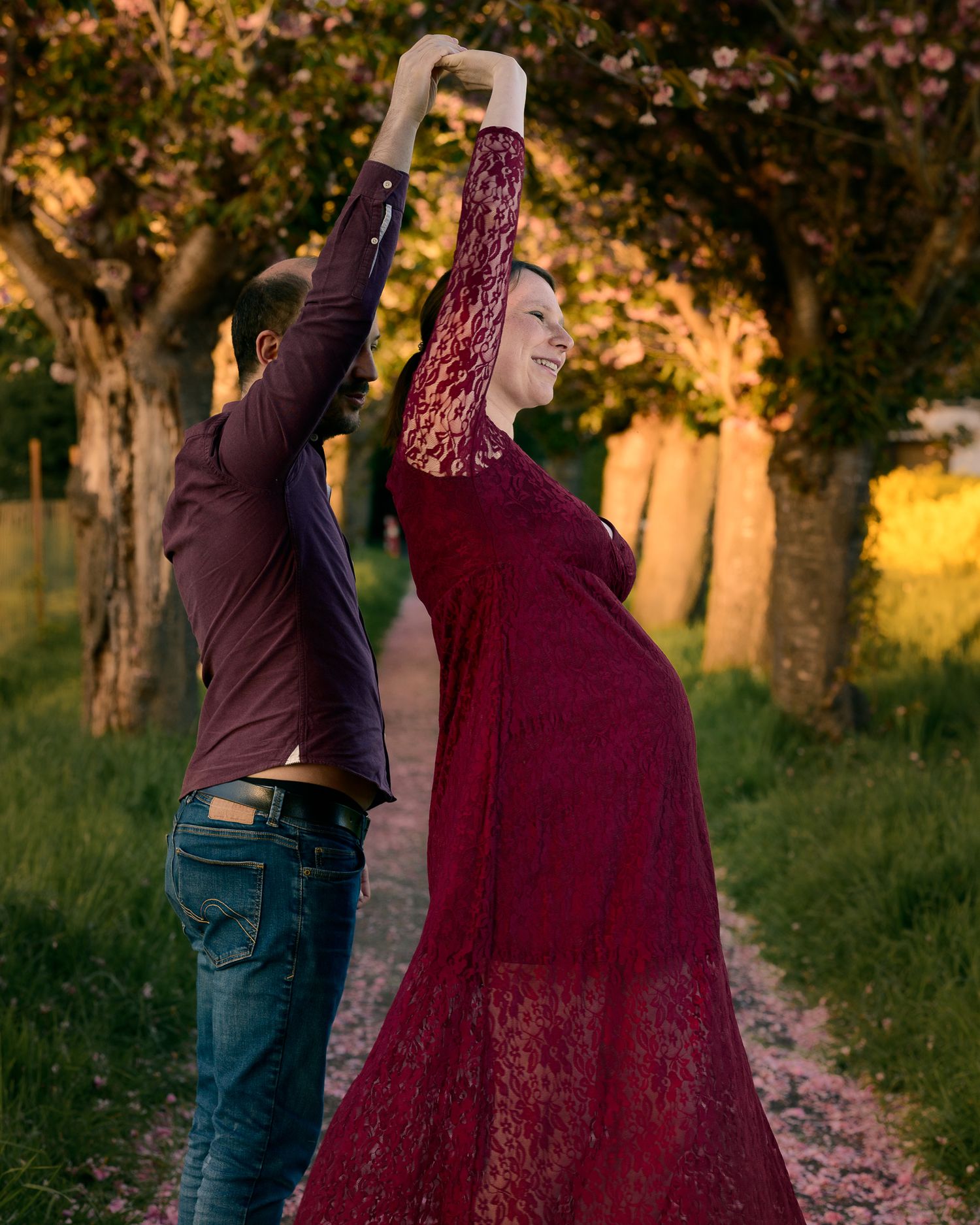 Couple dancing outdoors during golden hour, pregnant woman in burgundy lace gown twirling on a tree-lined path.