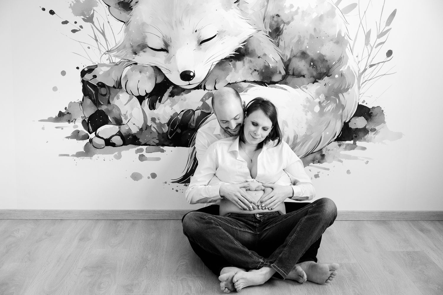 Black and white photo of expecting couple sitting on floor, hands on baby bump, with watercolor fox mural behind them.