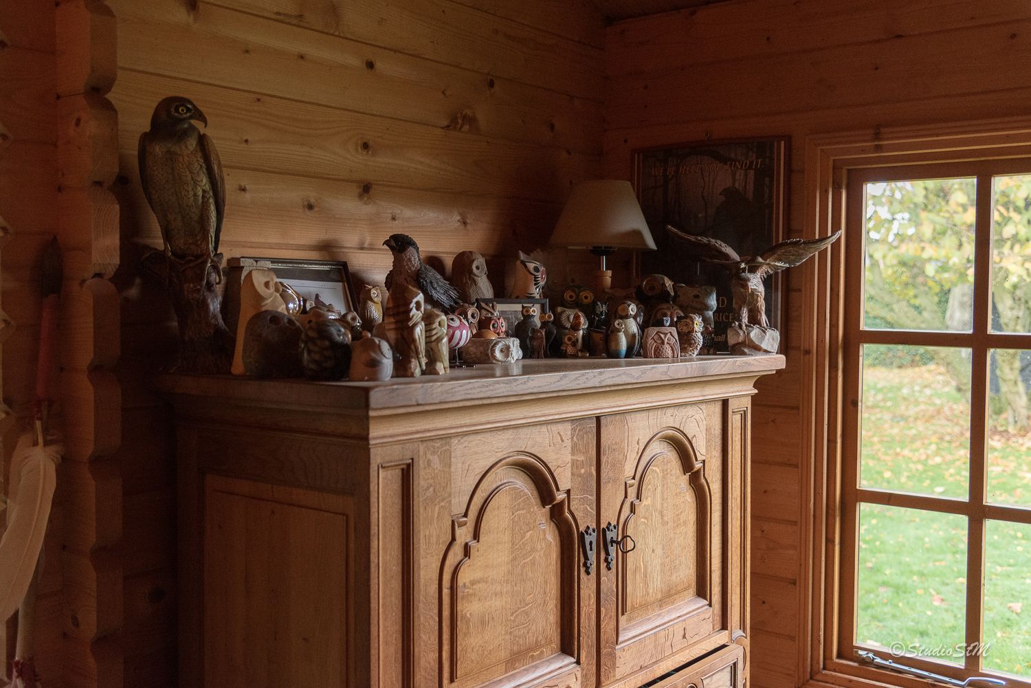 Rustic log cabin interior with wooden armoire topped with bird figurines and animal sculptures near a window.
