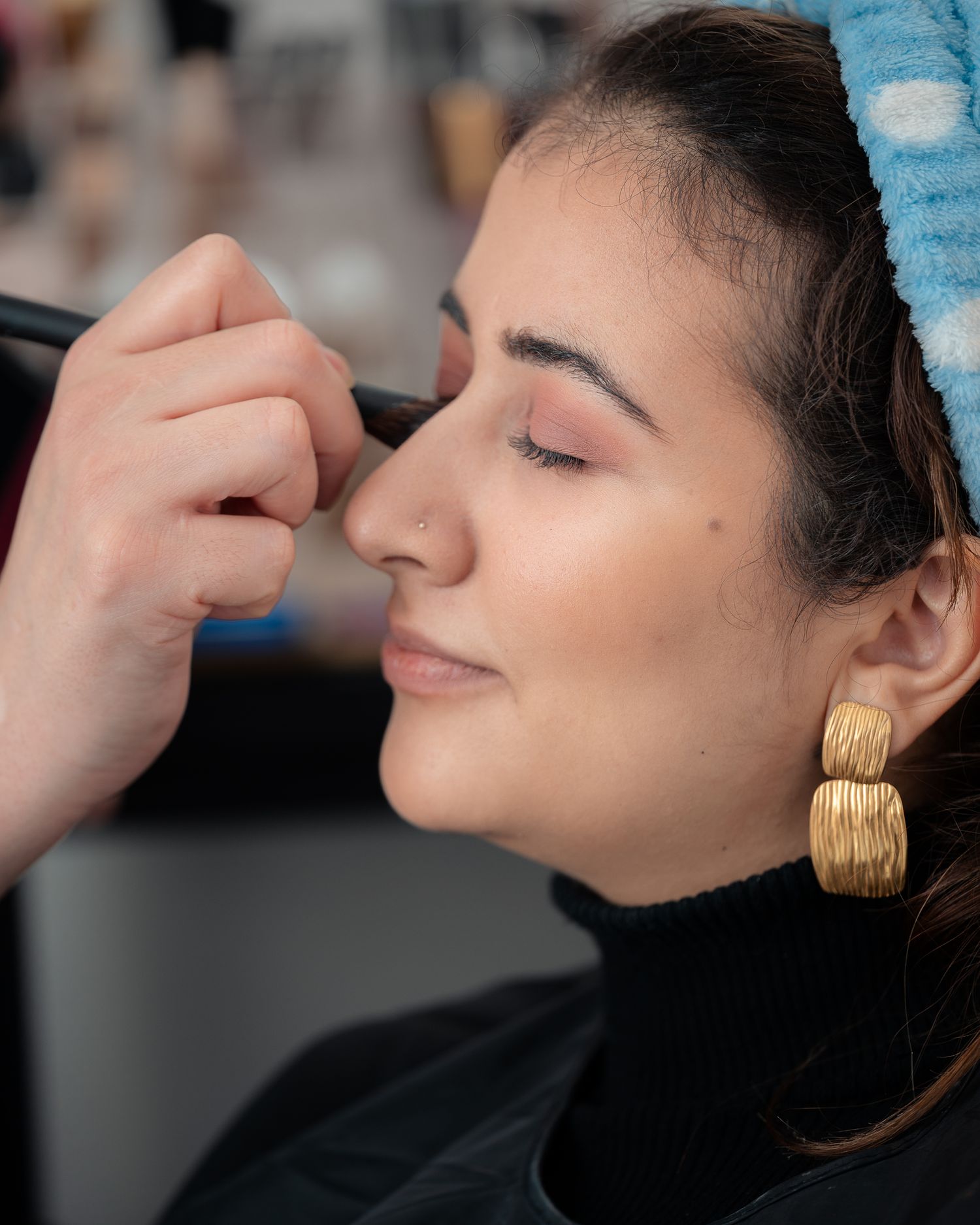 Woman getting makeup applied with a brush, wearing gold earrings and blue headband, eyes closed.