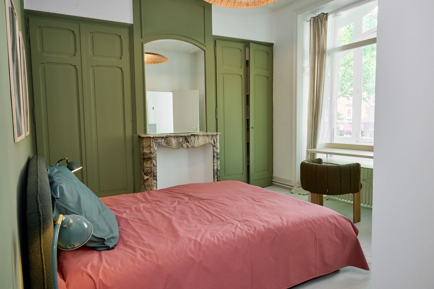 Elegant bedroom with olive green paneled walls, pink bedding, decorative fireplace, and natural light from a large window.