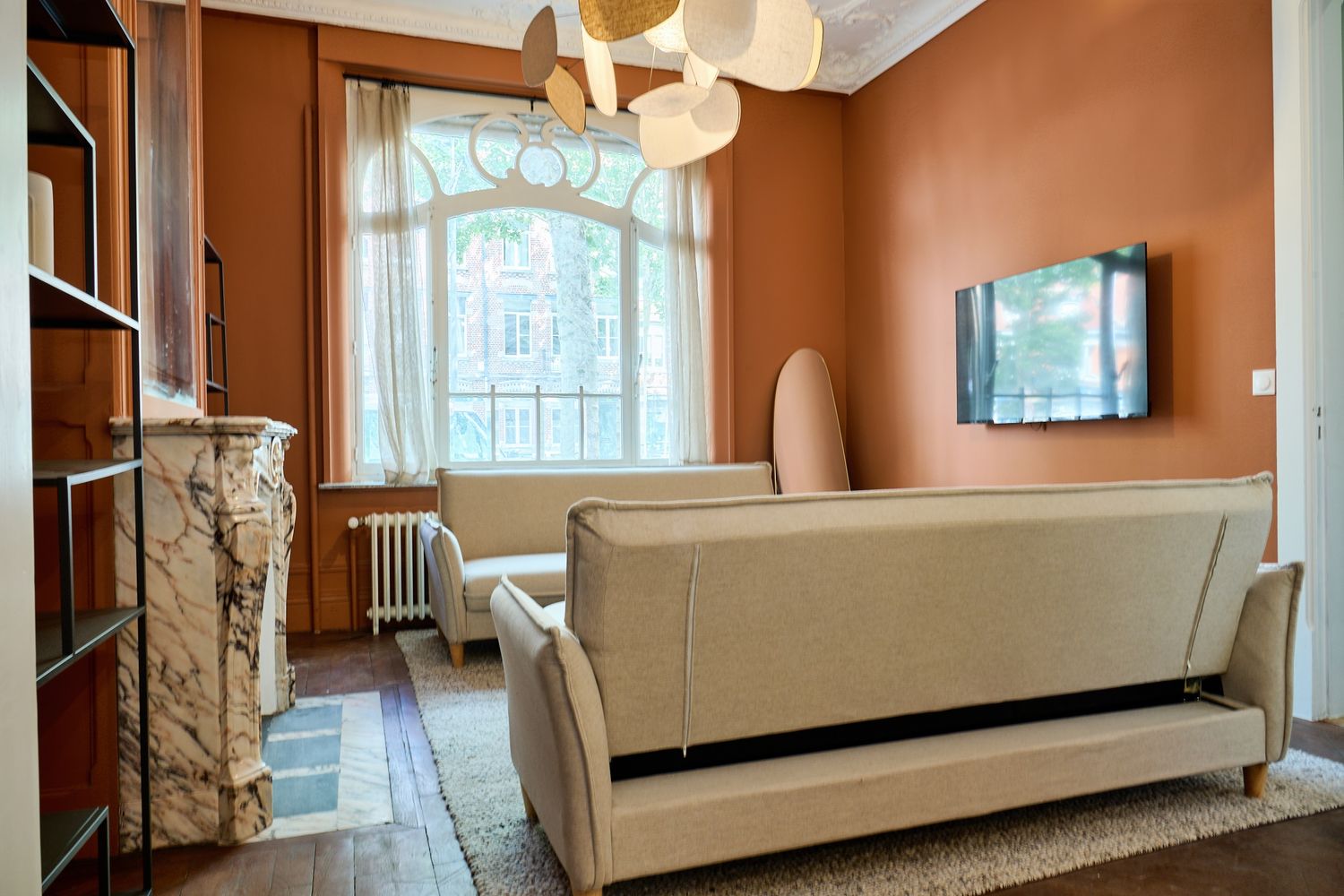 Cozy living room with terracotta walls, beige sofa, arched window, wall-mounted TV, and elegant chandelier on hardwood floors.