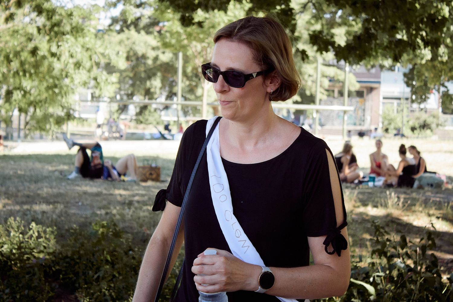 Woman in black top and sunglasses carries a white bag while walking through a sunny park.