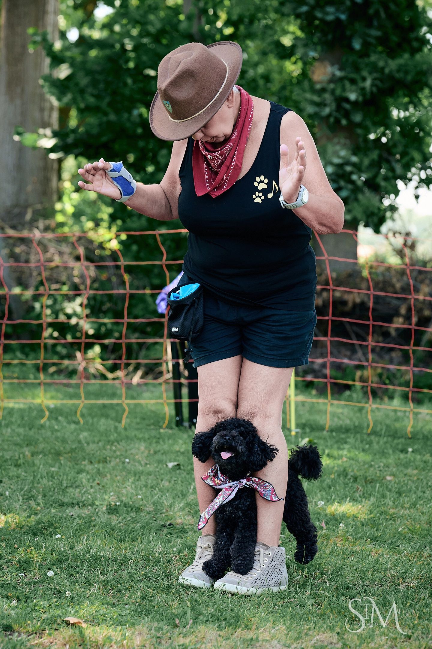 Woman in cowboy hat and red bandana training a small black poodle wearing a bandana on green grass.