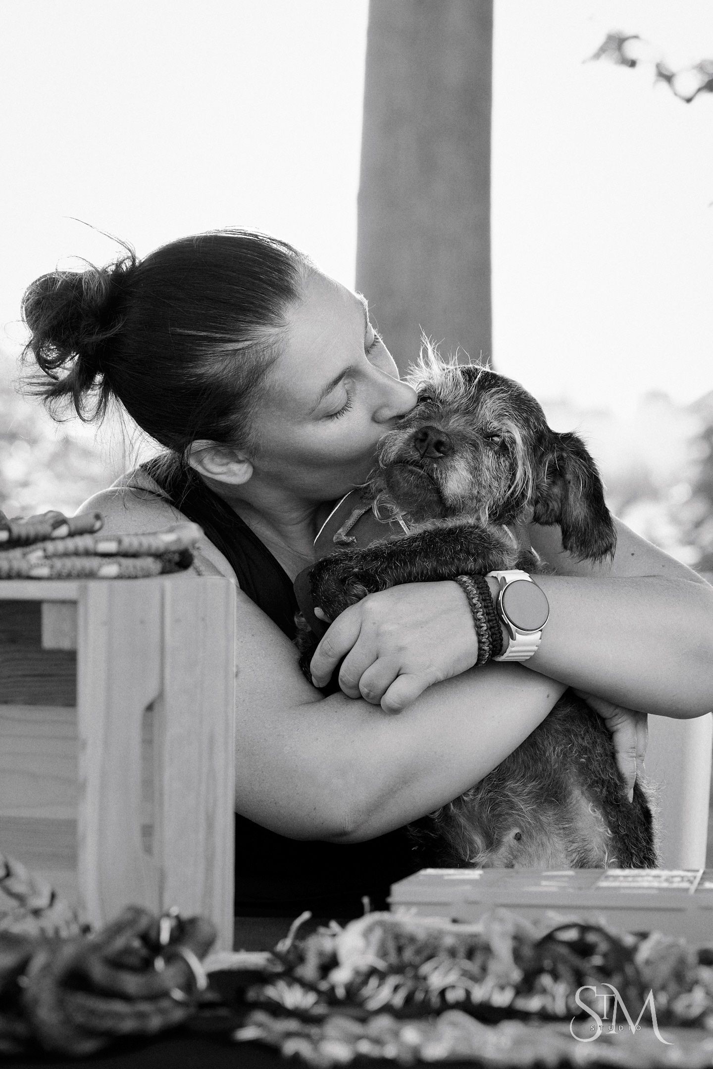 Black and white photo of a woman kissing her small scruffy dog while holding it in her arms outdoors.