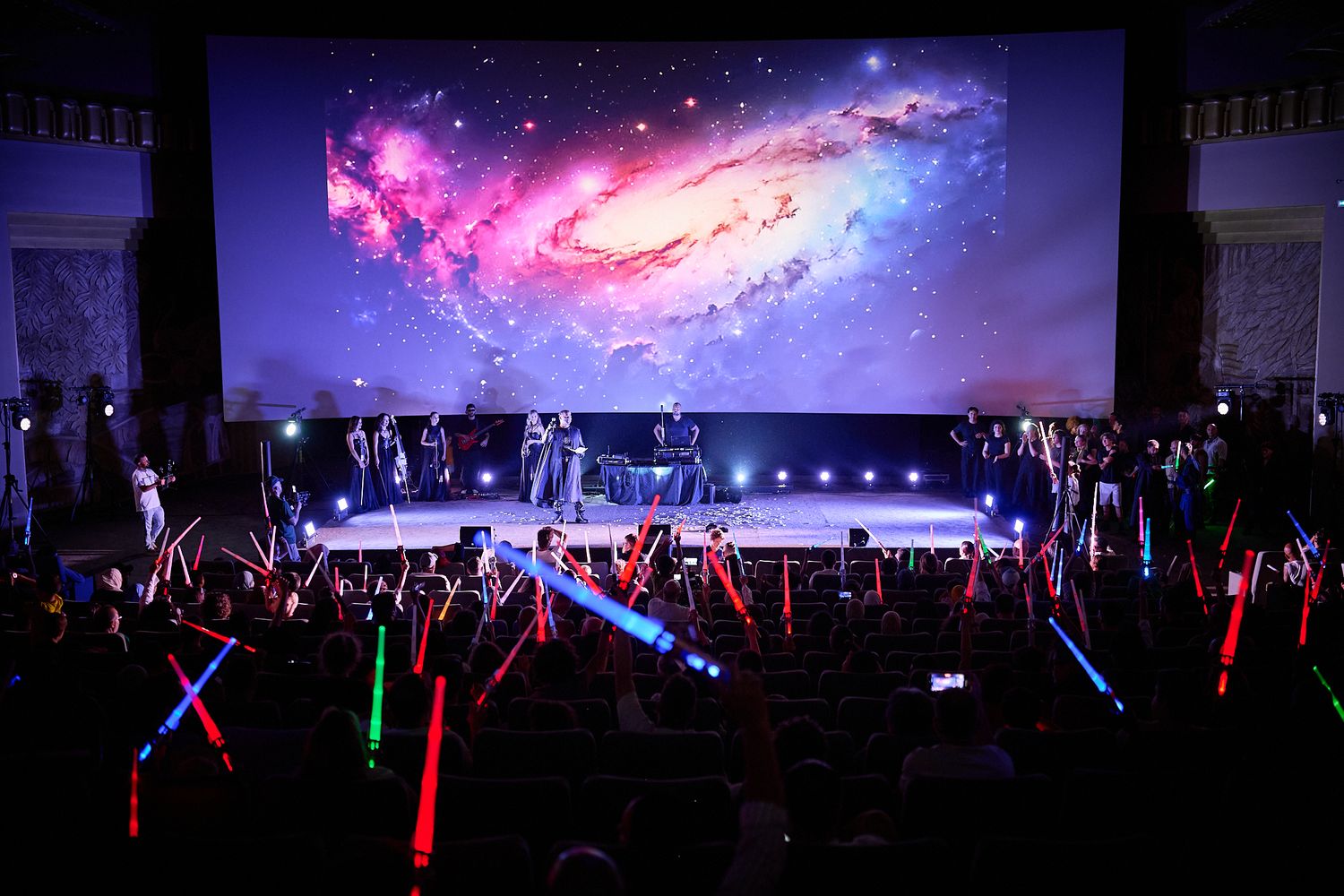 Audience waves colorful lightsabers at a Star Wars themed event with a galaxy projected on a large screen.