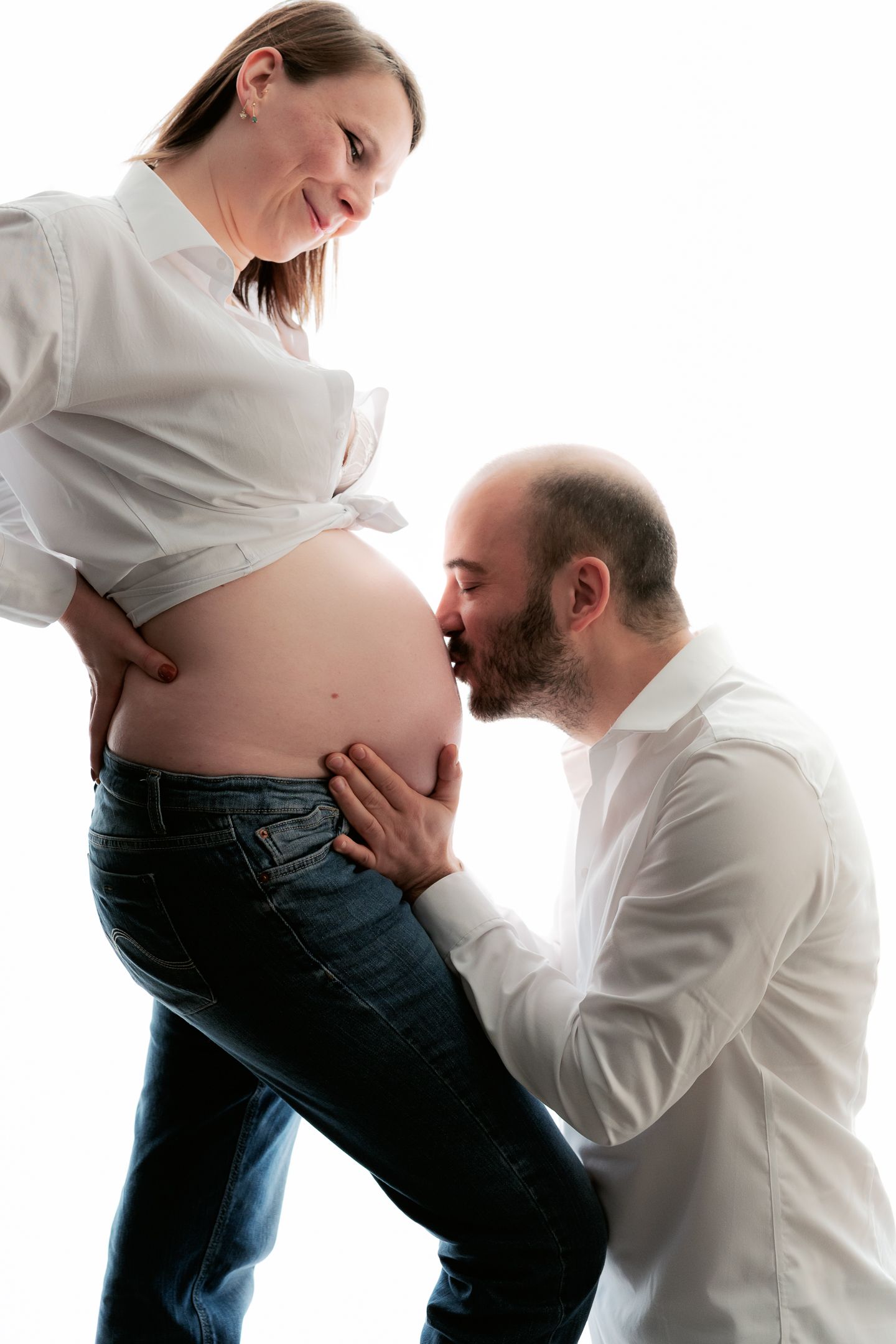 Man kissing pregnant woman's baby bump during maternity photo shoot on white background.