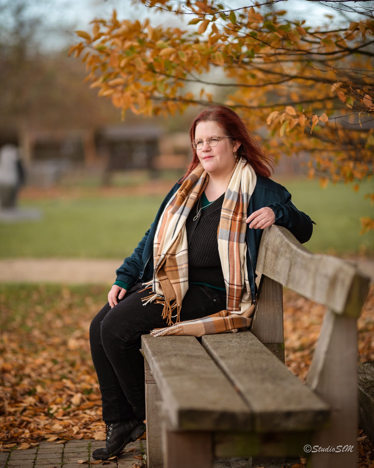 Red-haired woman in teal jacket sitting on wooden park bench surrounded by colorful autumn leaves.