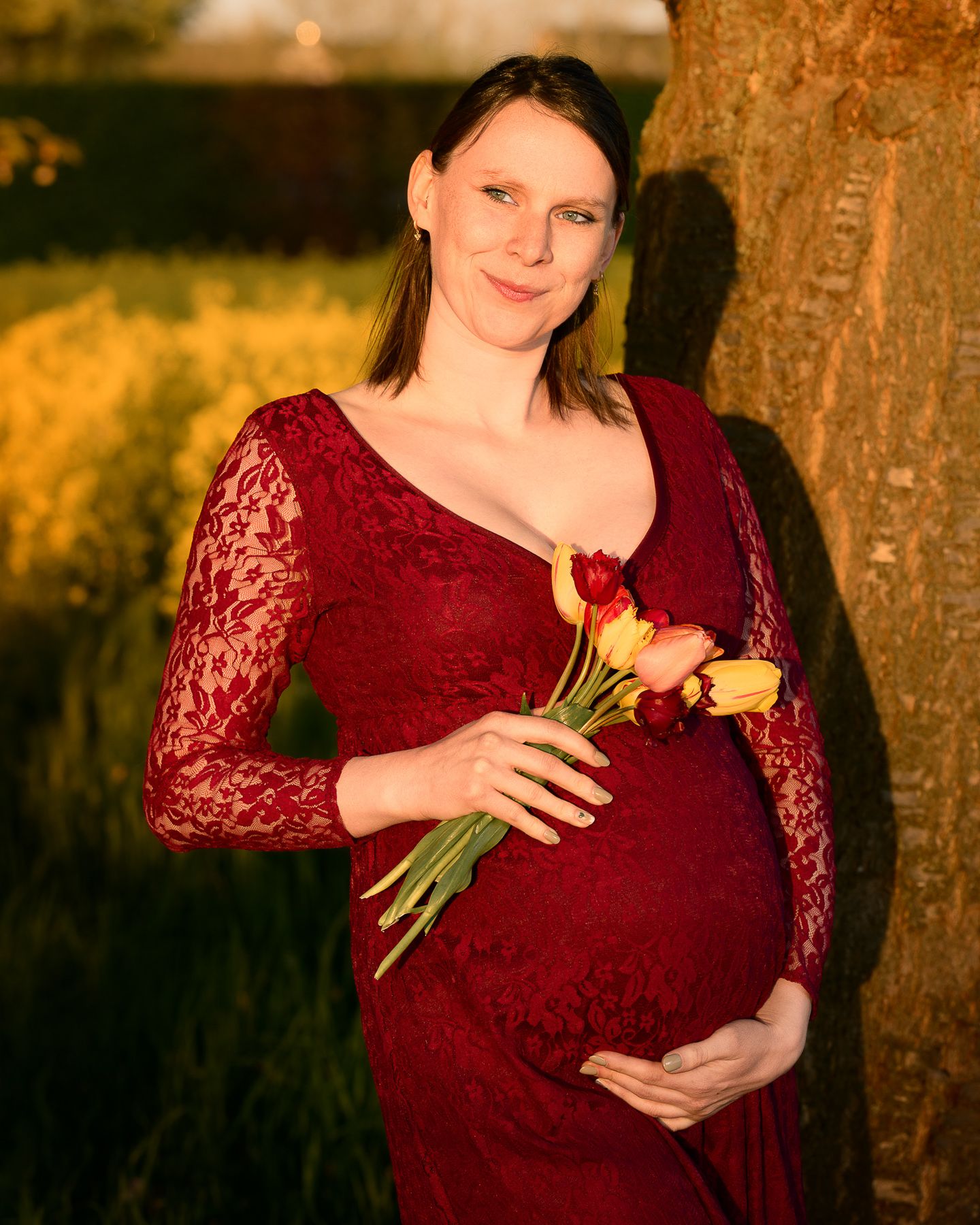 Pregnant woman in red lace dress holding tulips while leaning against tree in golden light.