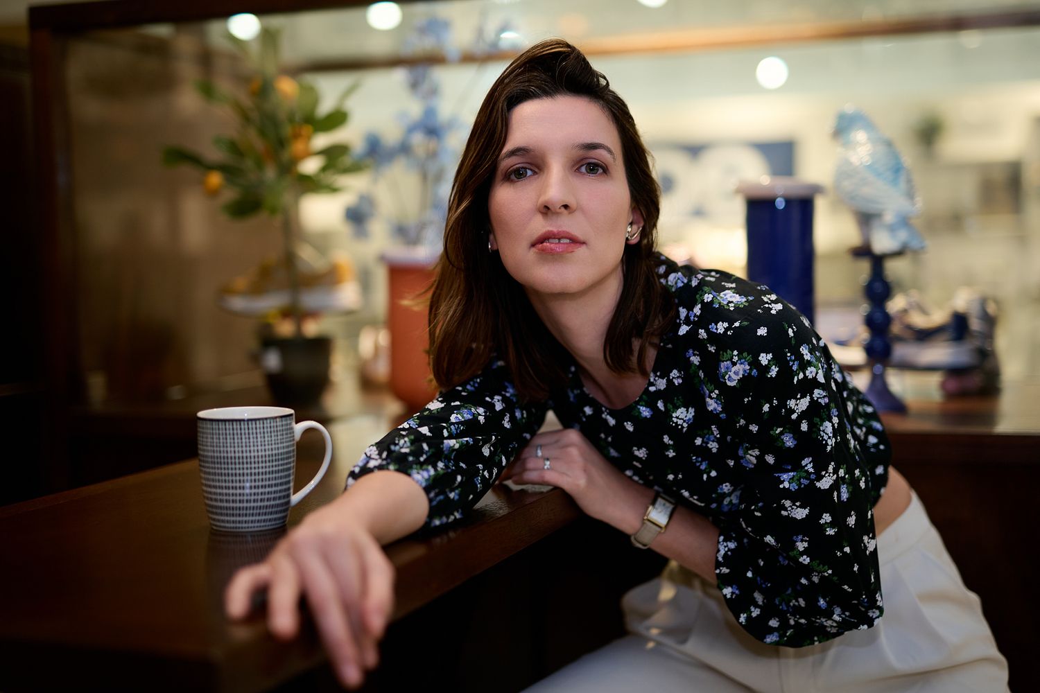 Thoughtful brunette woman sitting at table with coffee mug in floral blouse indoors.