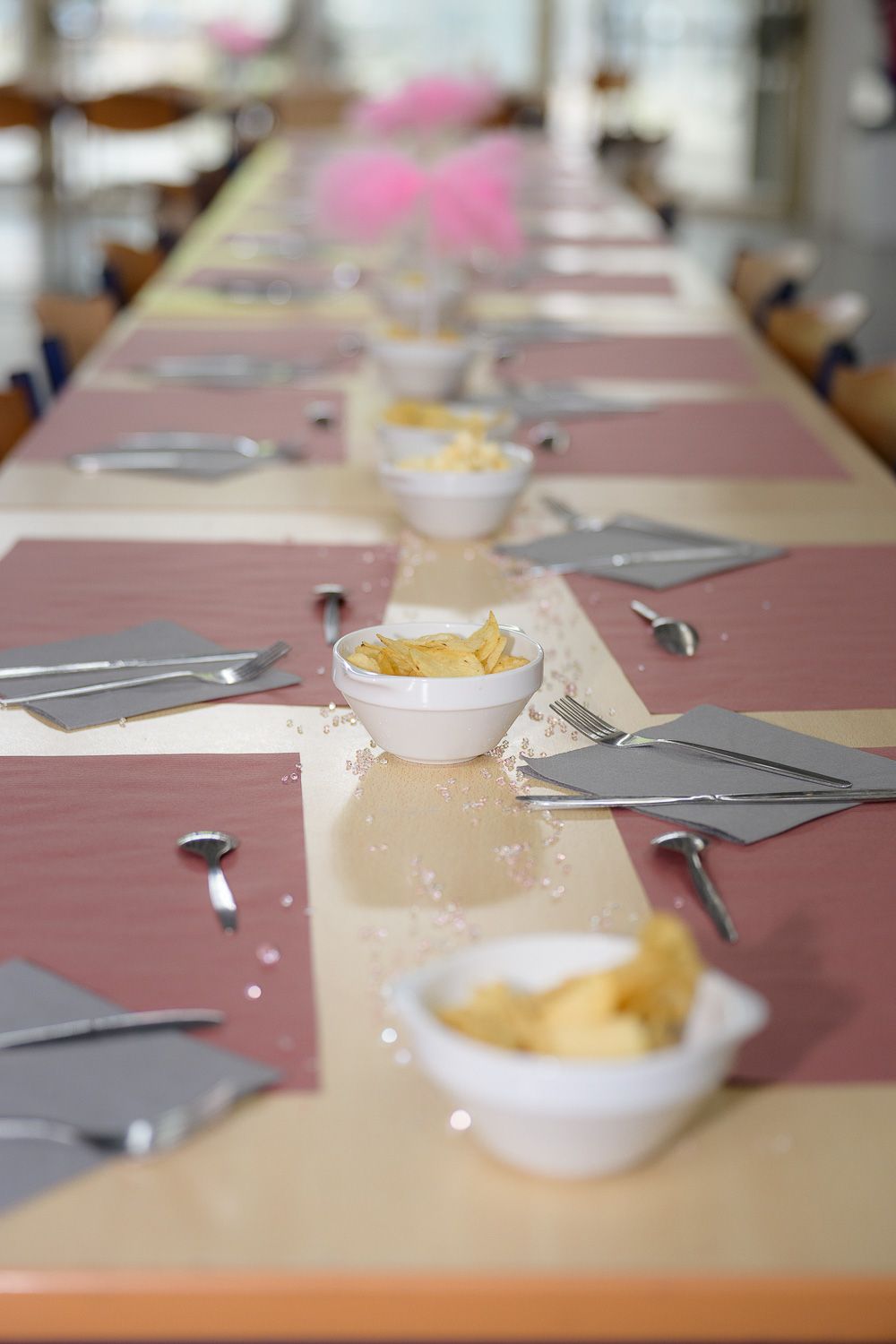 Long party table set with pink placemats, white bowls of chips, silverware, and gray napkins for a celebration.