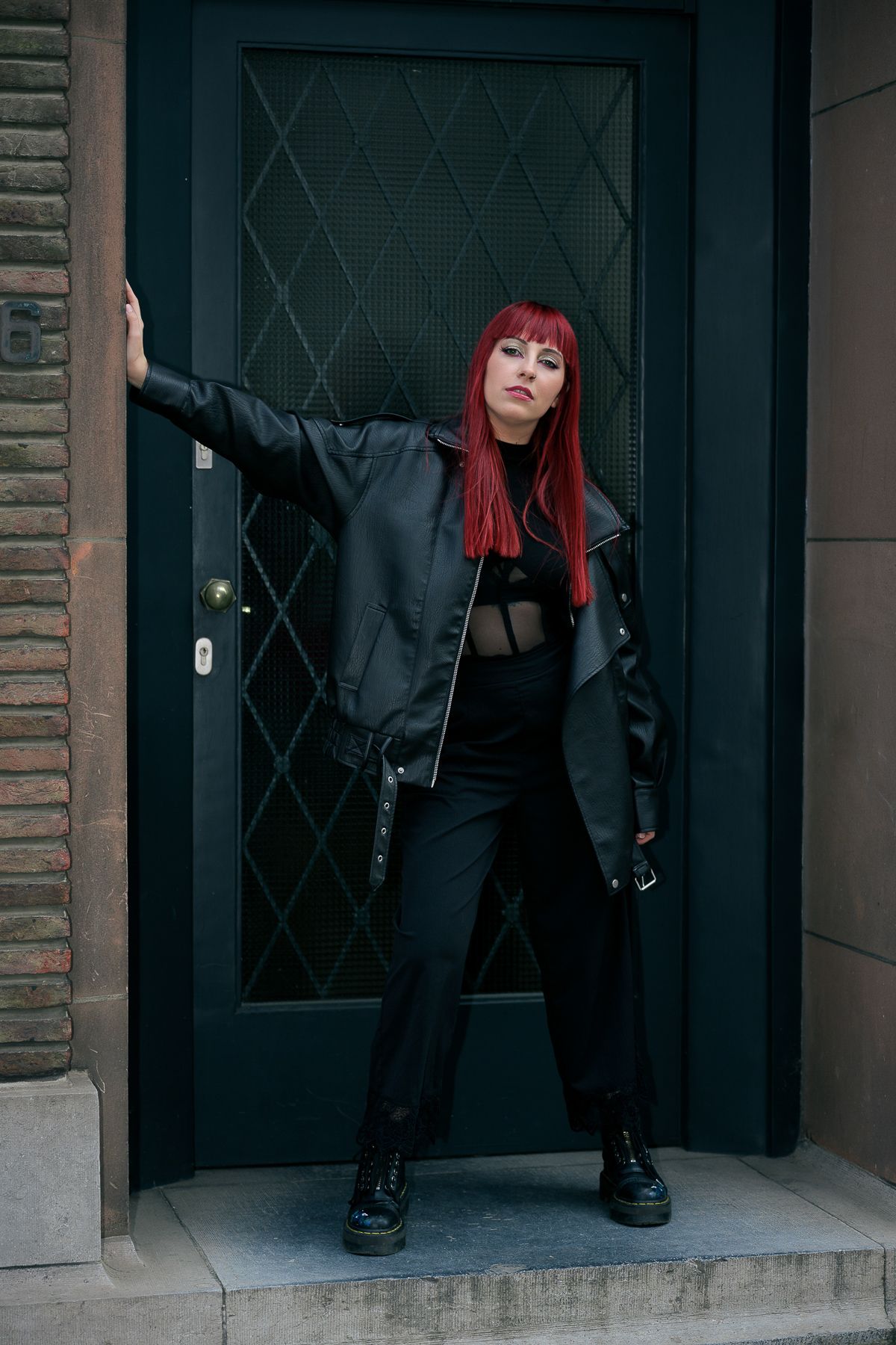 Woman with red hair in black leather jacket and trench coat posing against a dark green door.