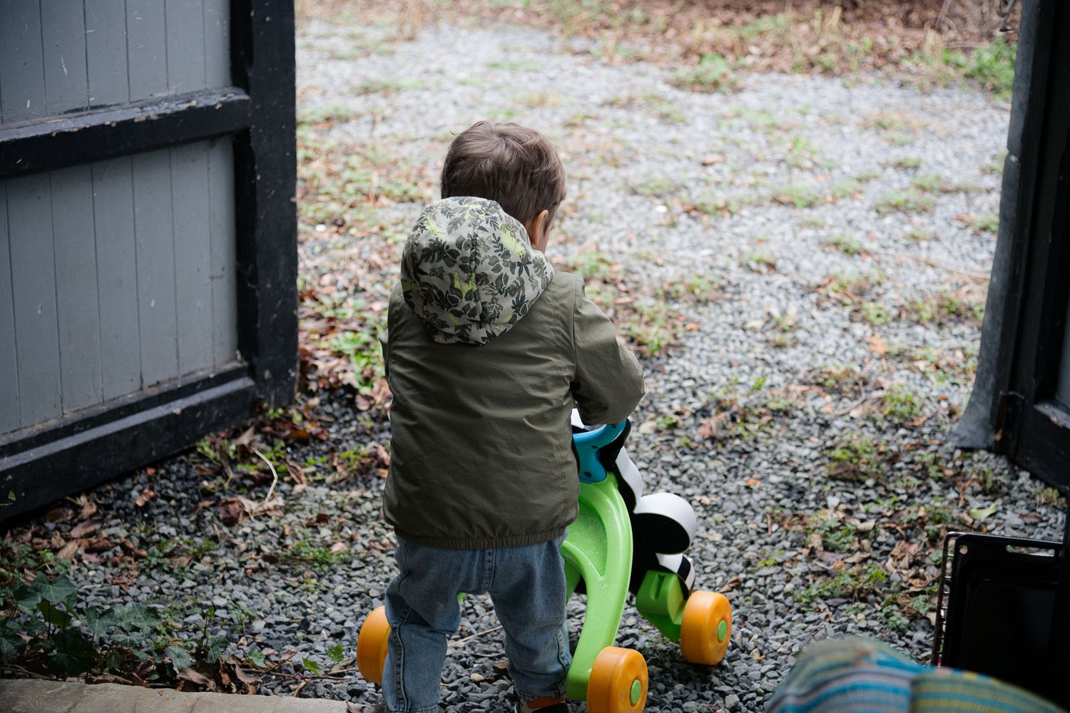 Toddler in green jacket riding a toy bike near an open door outside.