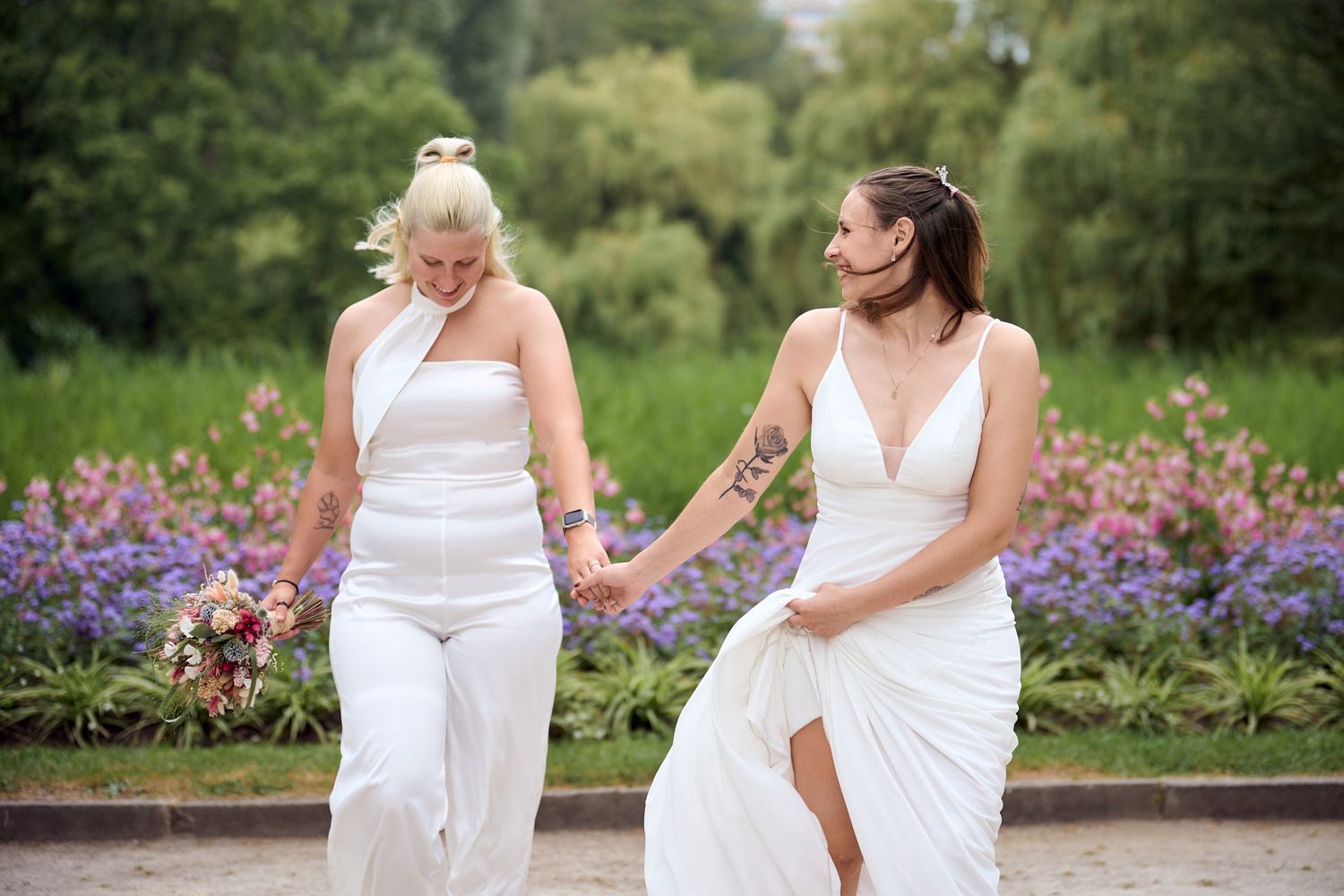 Two brides in white dresses hold hands walking past purple flowers in an outdoor wedding ceremony.