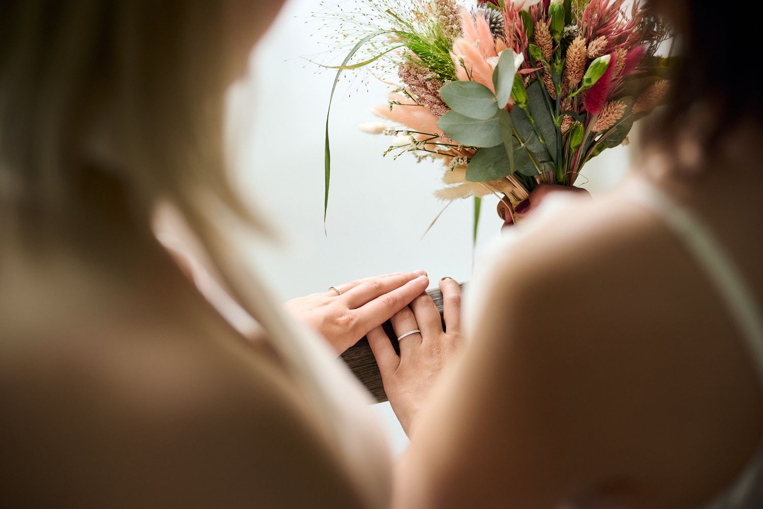 Two brides holding hands with a floral bouquet of pink flowers and greenery on their wedding day.