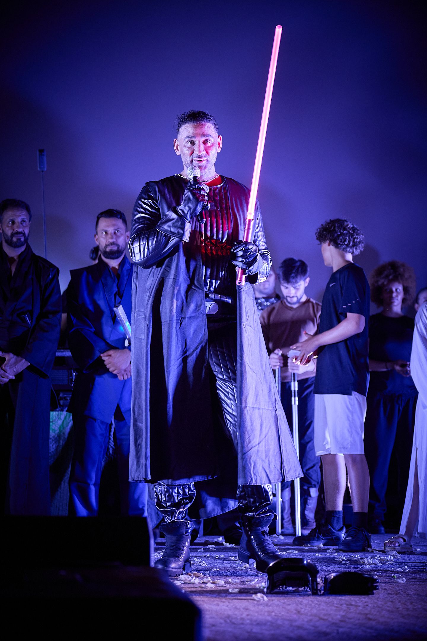 Man in dark Sith costume holds red lightsaber on stage at Star Wars themed event with audience behind him.