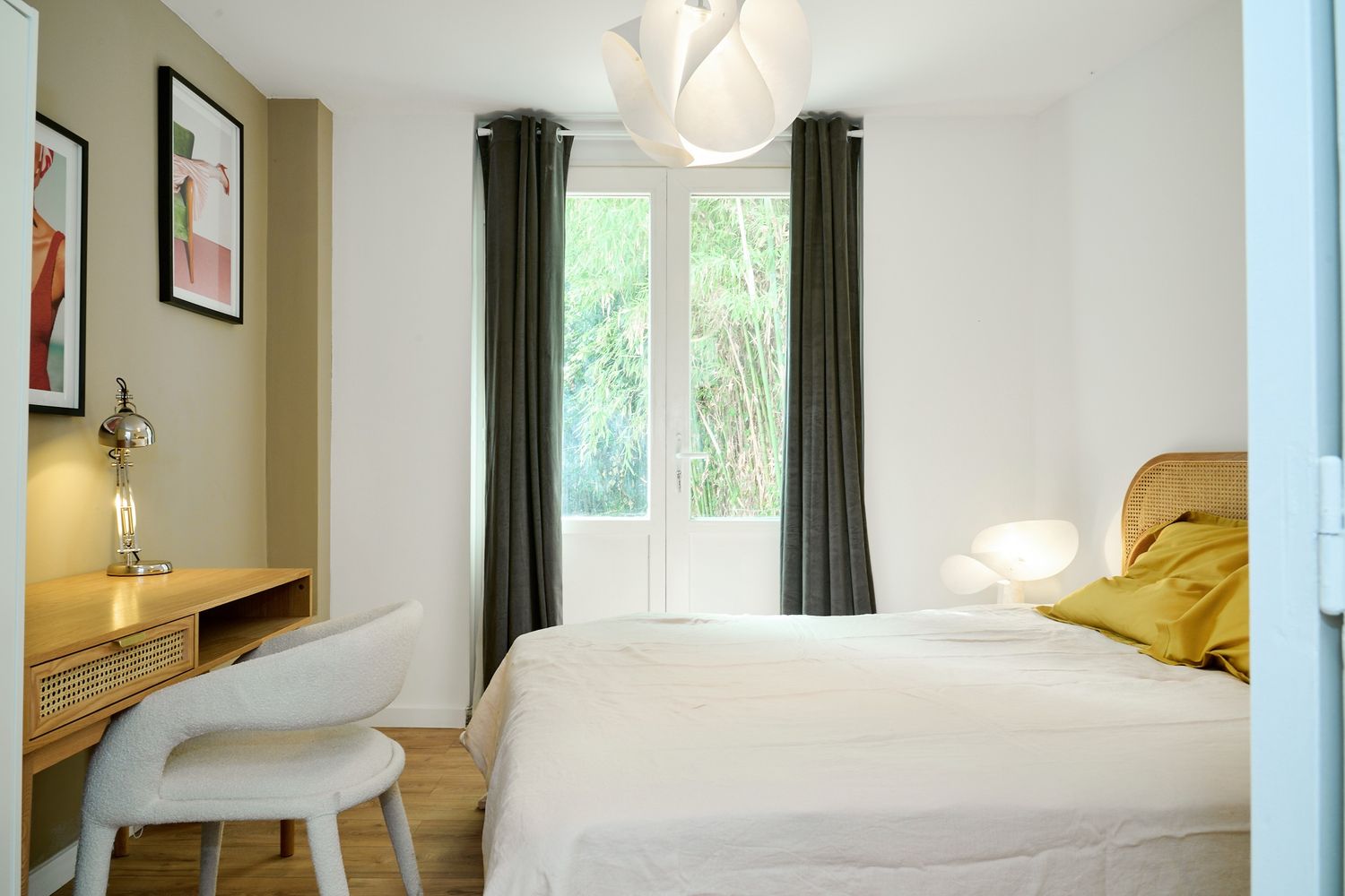 Bright modern bedroom with white bedding, mustard pillow, dark curtains, wooden desk, and white chair bathed in natural light.