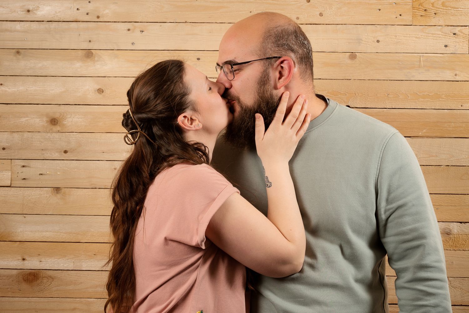 A couple sharing a romantic kiss against a wooden wall background, woman in pink top touching mans face.