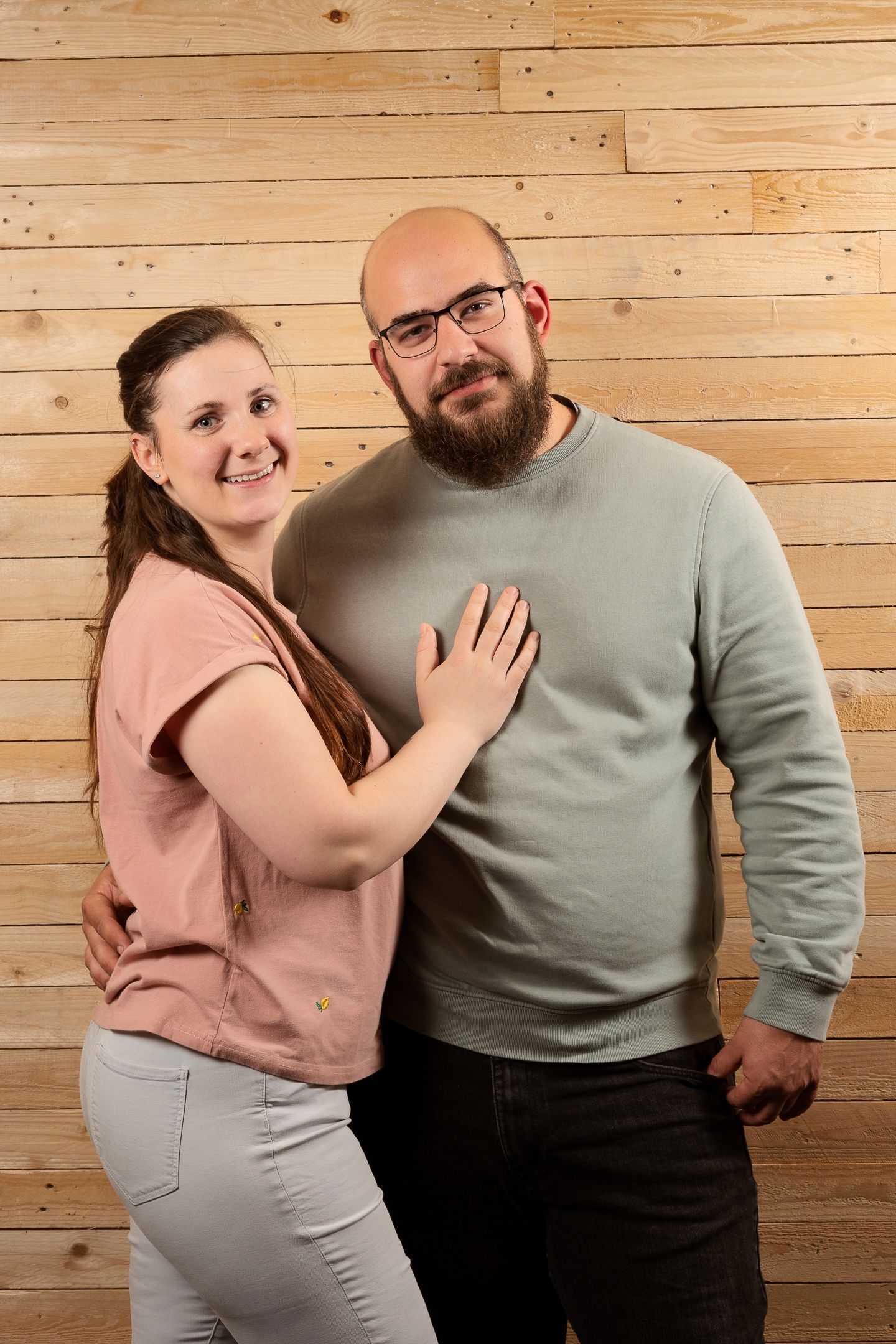 A smiling couple poses against a wooden wall, woman in pink top rests hand on bearded bespectacled mans chest.