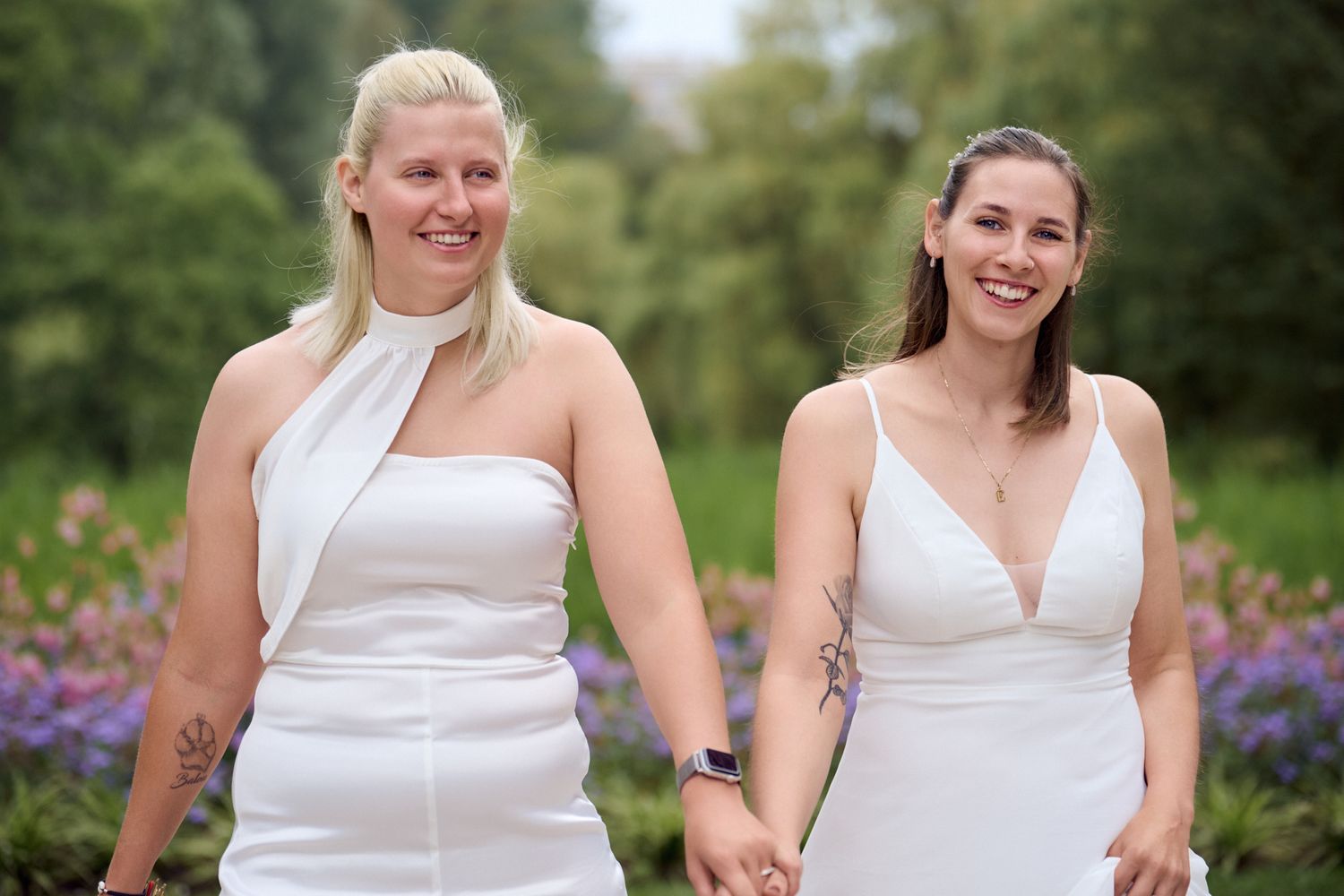 Two women in white wedding dresses hold hands and smile in a garden with purple flowers.