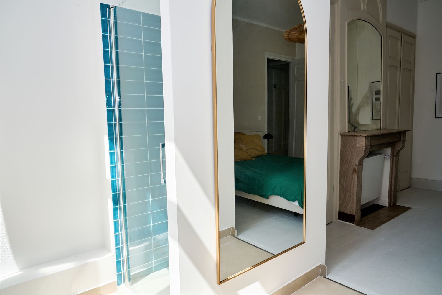Bright room with arched mirror reflecting a bedroom with teal bedding, beside a blue-tiled shower and stone fireplace.