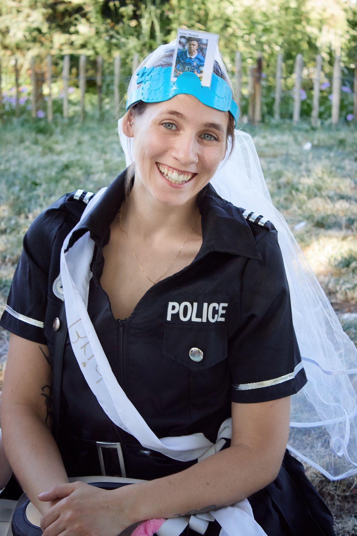 Smiling woman in police costume with bridal veil and photo hat accessory outdoors.