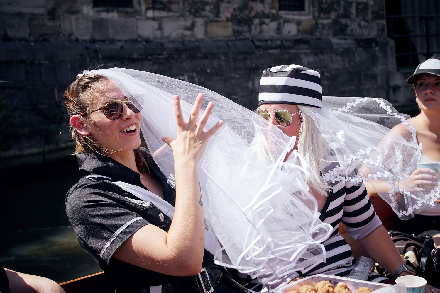 People in festive costumes, including a bridal veil, celebrating outdoors near a canal on a sunny day.