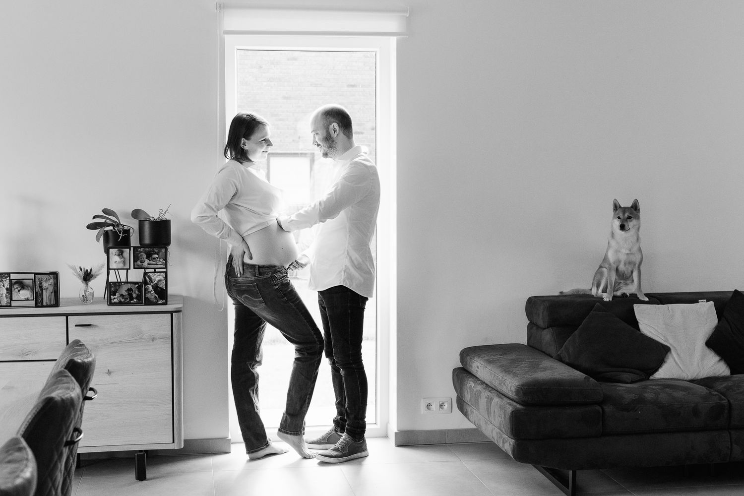 Black and white photo of a couple dancing in a doorway at home, with a cat on the sofa and another near a dresser.
