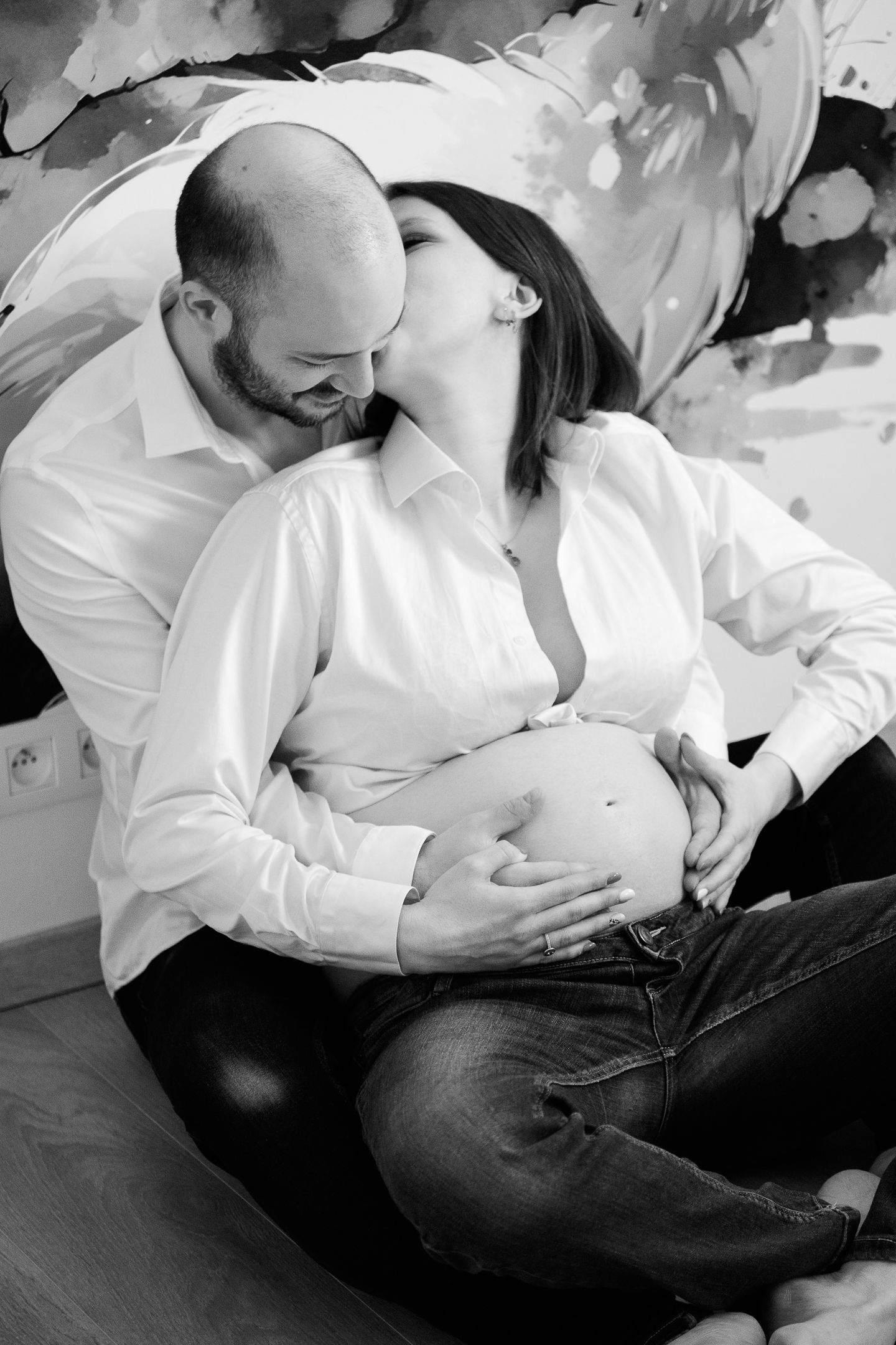 Black and white photo of a couple sharing a tender moment, with the woman kissing her partner while he cradles her pregnant belly.