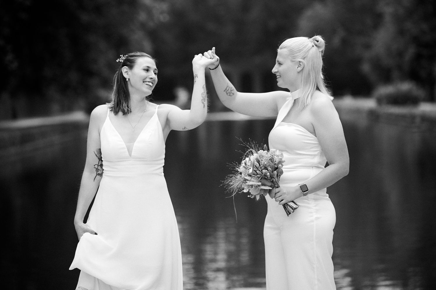 Two brides in white dresses hold hands and smile by a lake in a black and white wedding photo.