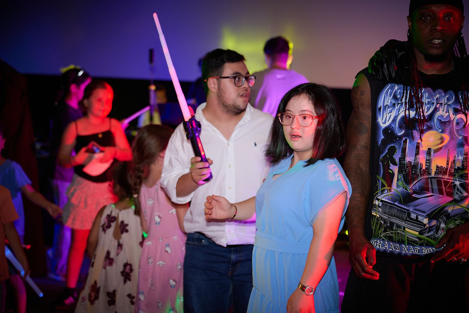 Young couple at a neon dance party, man holding glowing light saber, woman in blue dress wearing pink glasses.