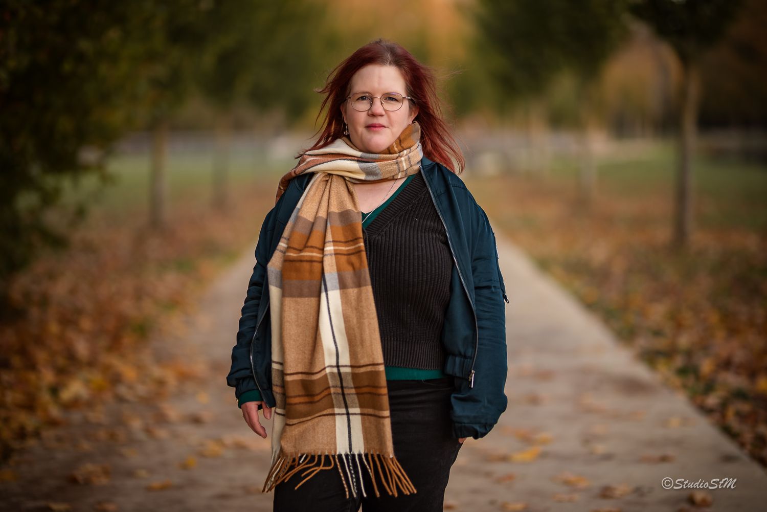 Red-haired woman in glasses and plaid scarf stands confidently on a tree-lined autumn path.