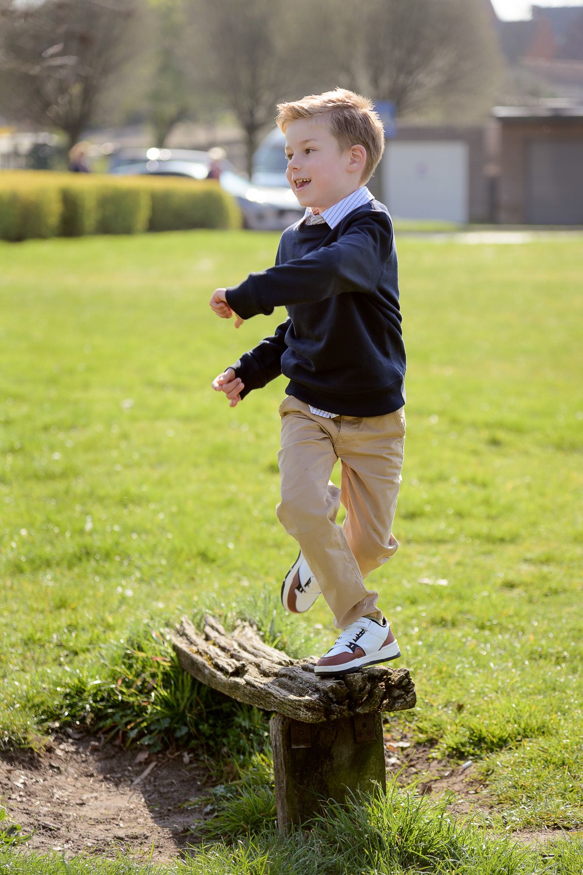 Young boy balancing on a tree stump in a sunny green park, wearing a navy sweater and khaki pants.
