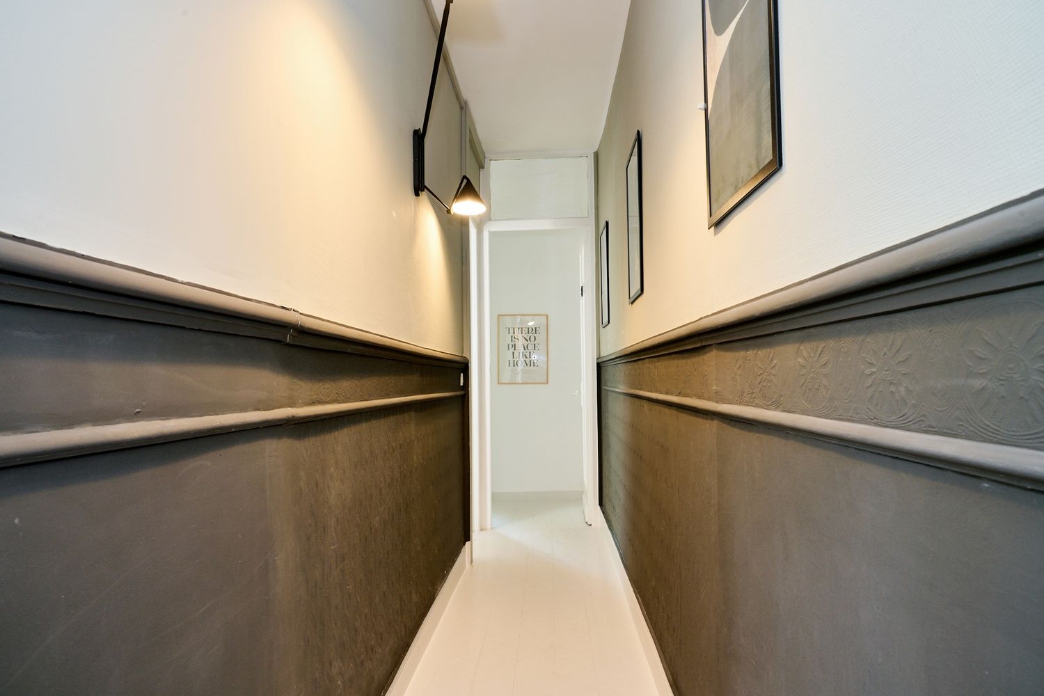 Narrow hallway with dark wainscoting, white walls, pendant light, and framed artwork leading to a bright doorway.