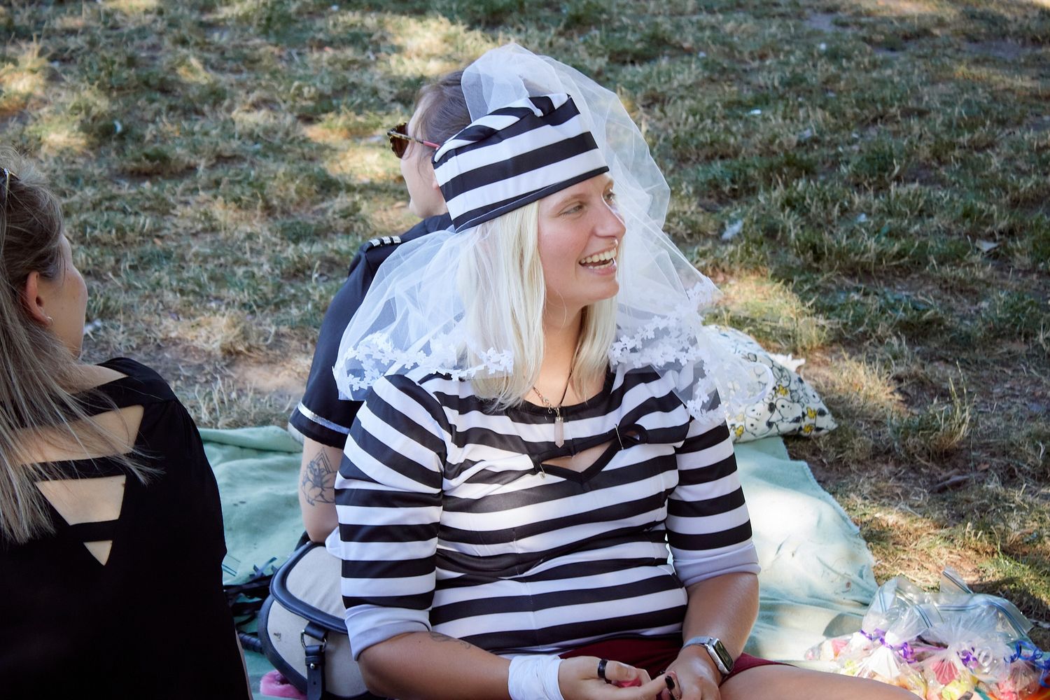 Smiling blonde woman in striped prisoner costume with white veil hat sitting outdoors at a themed party.