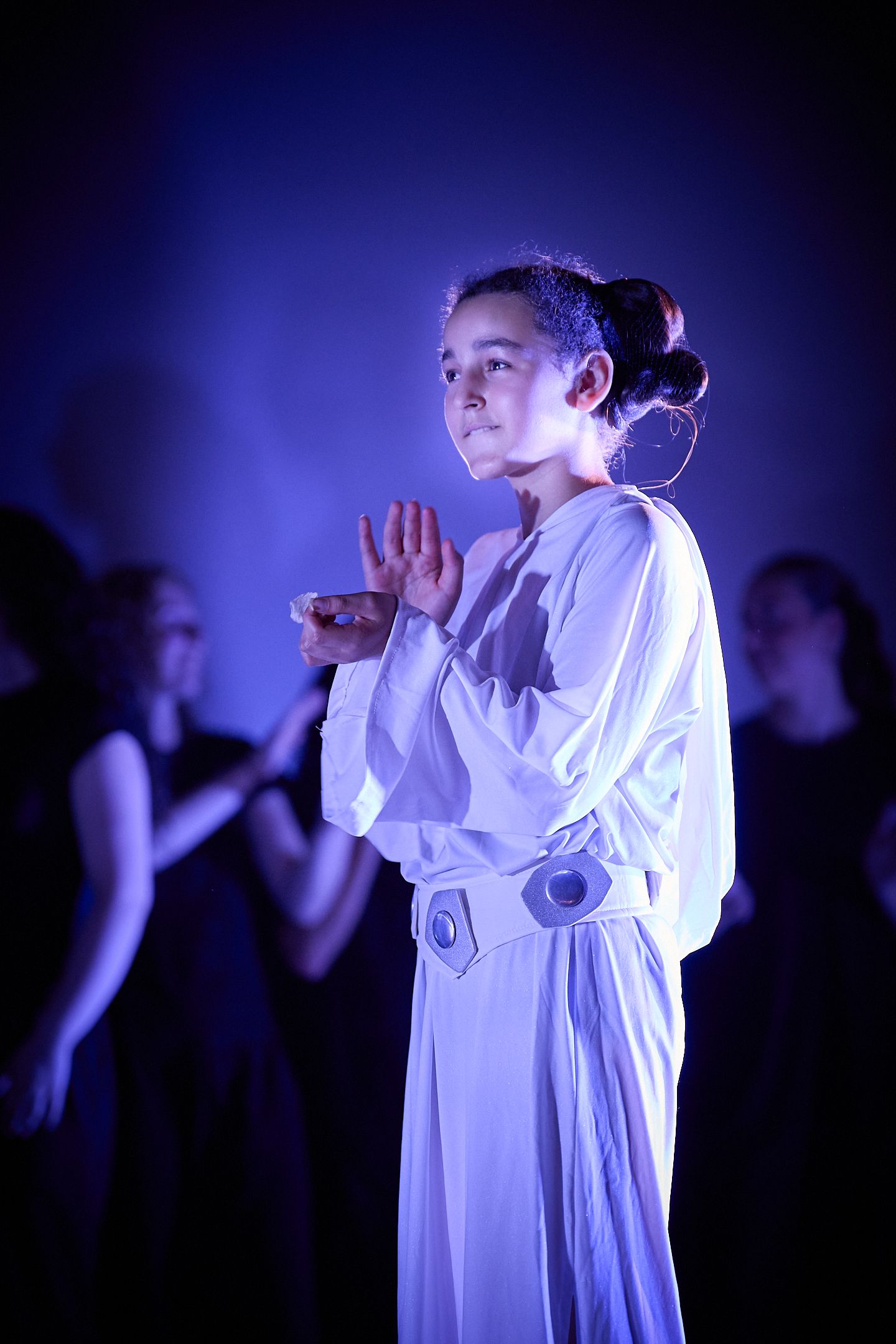 Young girl in white costume performs on stage under blue lighting during a theatrical production.