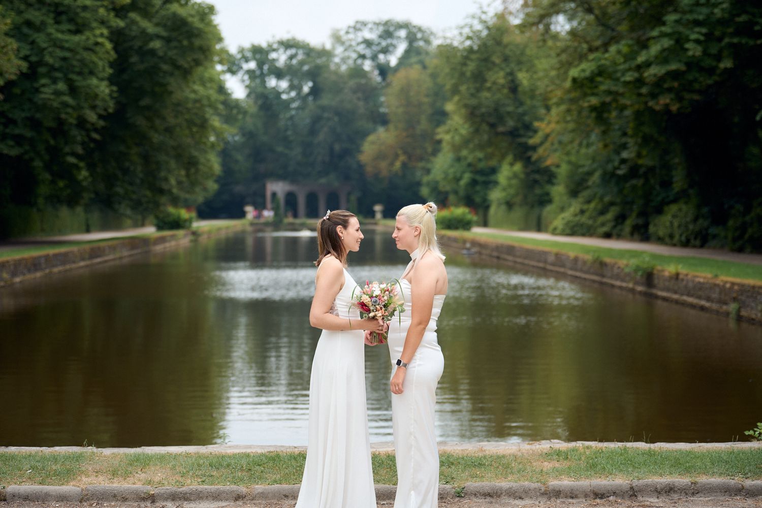 Two brides in white attire hold bouquet by a serene garden canal surrounded by lush green trees.
