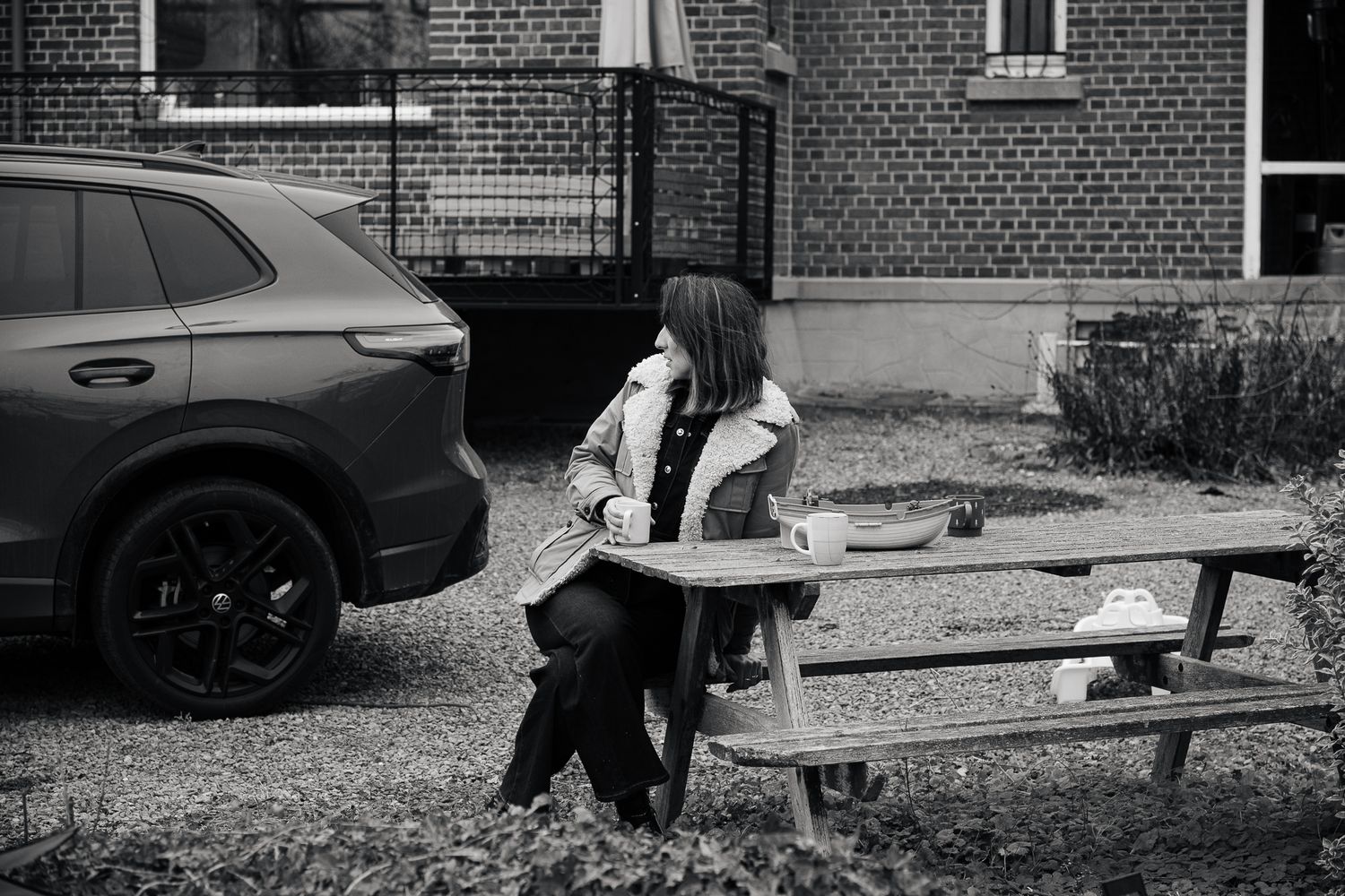 Black and white photo of a woman sitting alone at an outdoor picnic table next to a parked SUV.