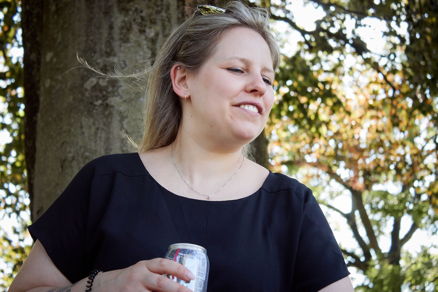 Woman in black top holding a beverage can outdoors, smiling near a large tree on a sunny day.